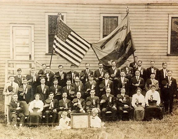 A black-and-white photo of a large group of men and a few women gathered outdoors in front of a wooden building, holding American flags and other banners. Some individuals are seated, others are standing, and a few children are seated in front. Many are dressed in suits and formal attire.