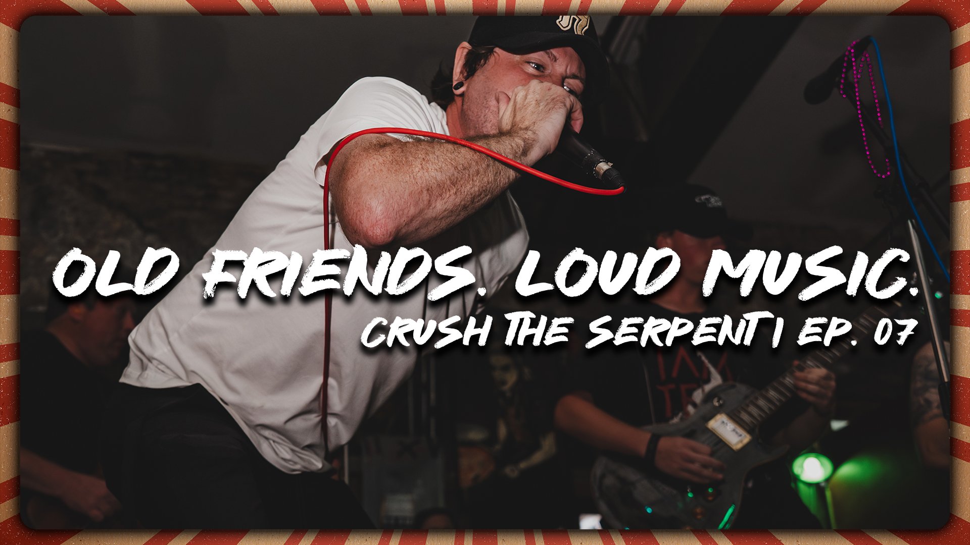 A man singing into a microphone during a live music performance with others playing instruments in the background, in a dimly lit setting.