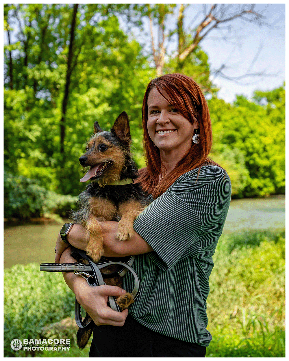 A woman with red hair smiling and holding a small, brown and black dog outdoors near a river with green trees in the background.