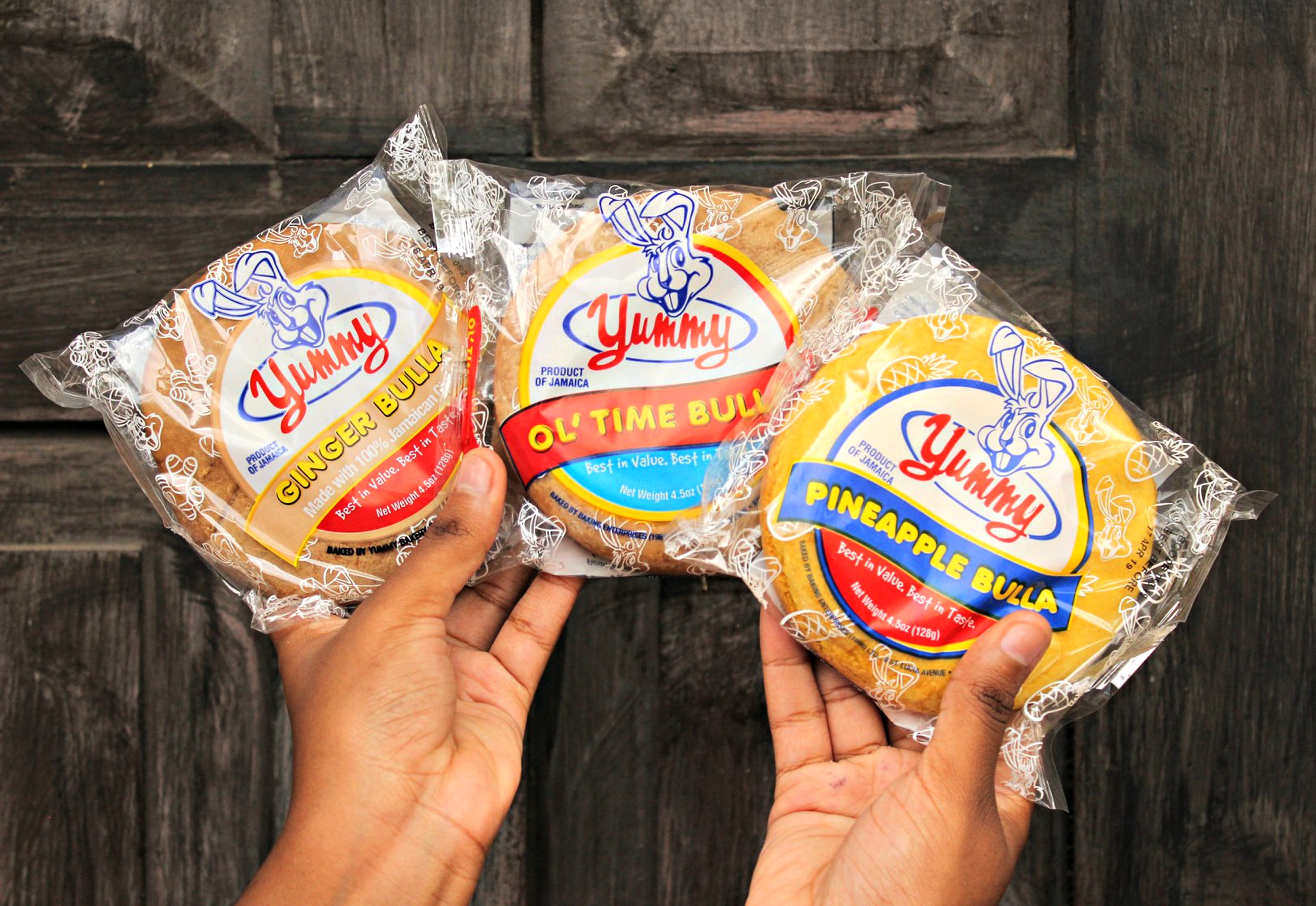 Three packages of Yummy Bulla baked bread flavors: ginger, ol' time, and pineapple, held by two hands against a dark wooden background.