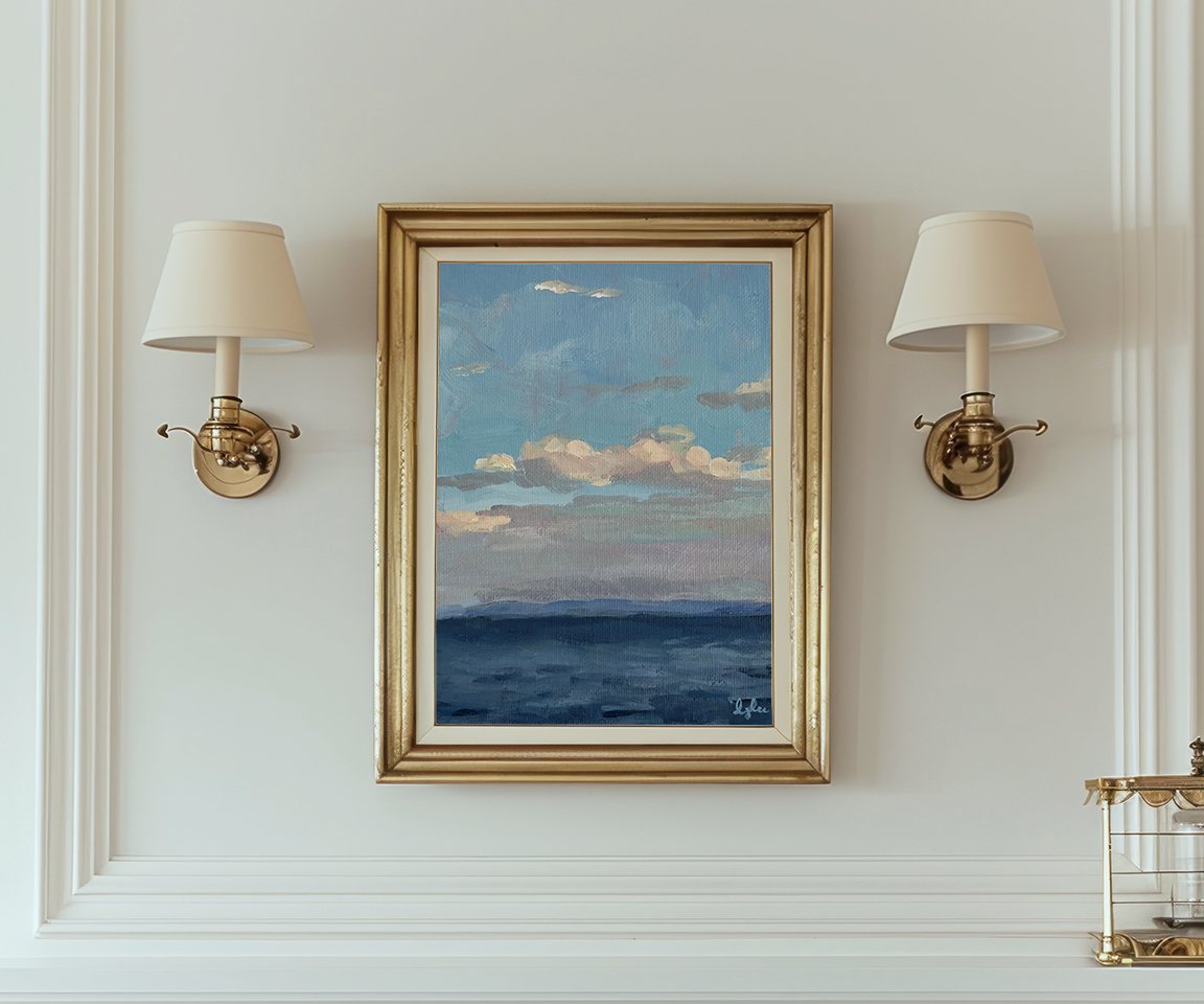 two scones either side of a gold framed print of a painting of clouds in blue sky over ocean