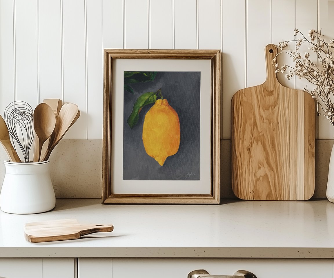 matted and framed print of painting of lemon with leaf on gray background sitting on kitchen counter with kitchen items