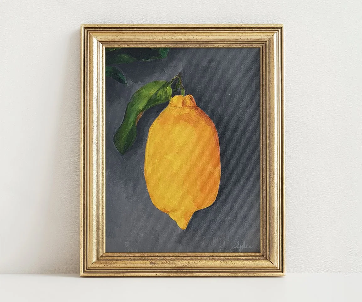 gold framed painting of lemon with leaf on gray background sitting on white wall