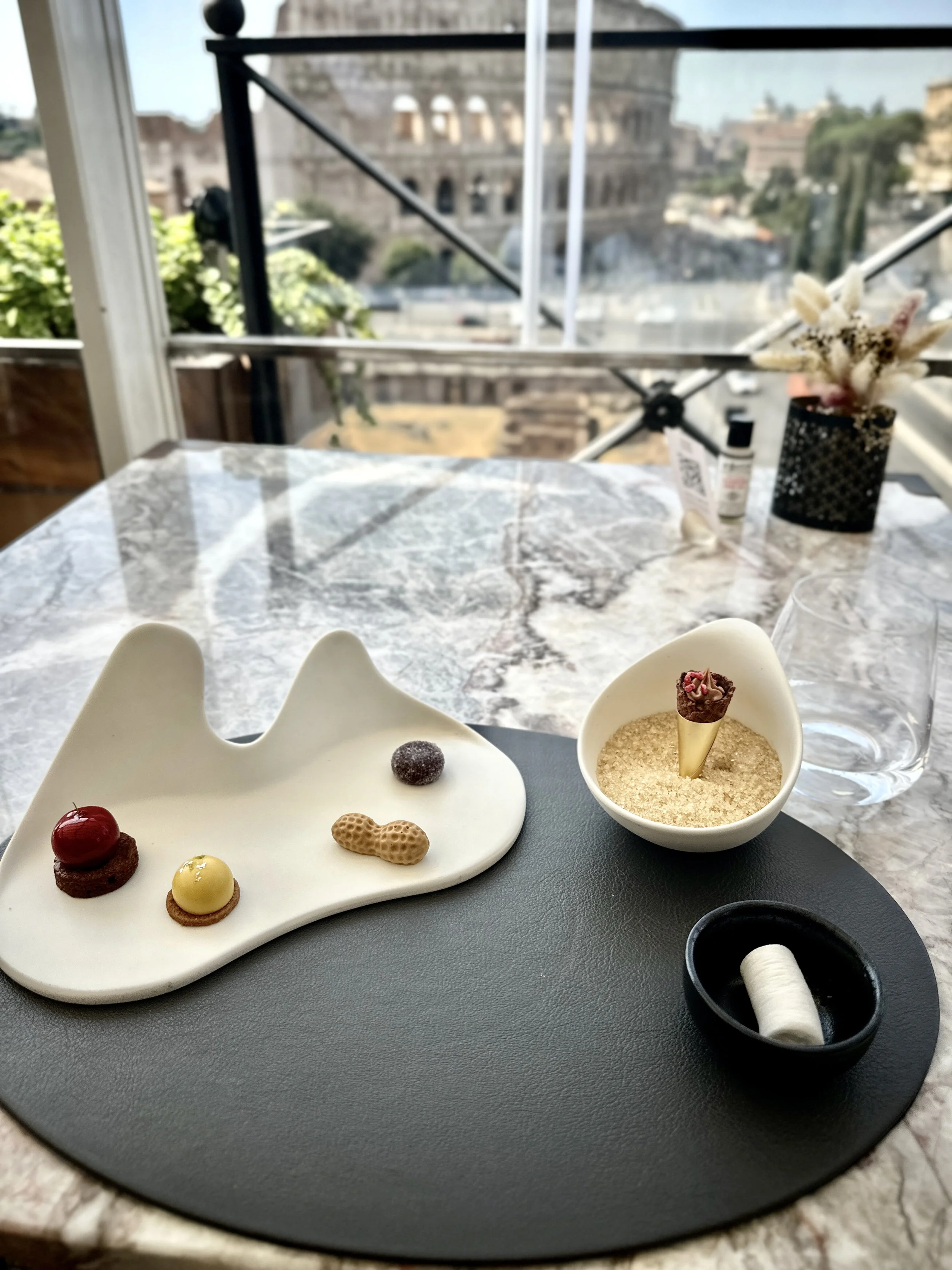 A marble table displaying three small pastries with decorative toppings on a white ceramic tray, a small cone-shaped dessert in a white bowl with crushed topping, and a black dish with a white spool of thread, set in a bright room with large windows and a city view.