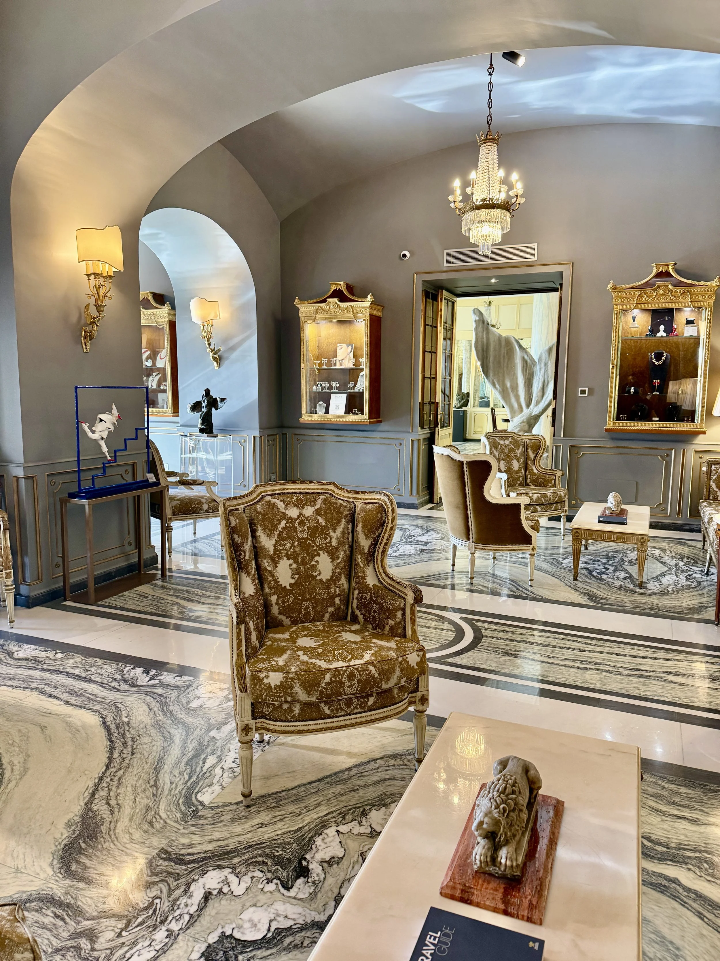 Luxurious interior of a room with ornate furniture, marble flooring, display cabinets with jewelry, and a chandelier hanging from a curved ceiling.