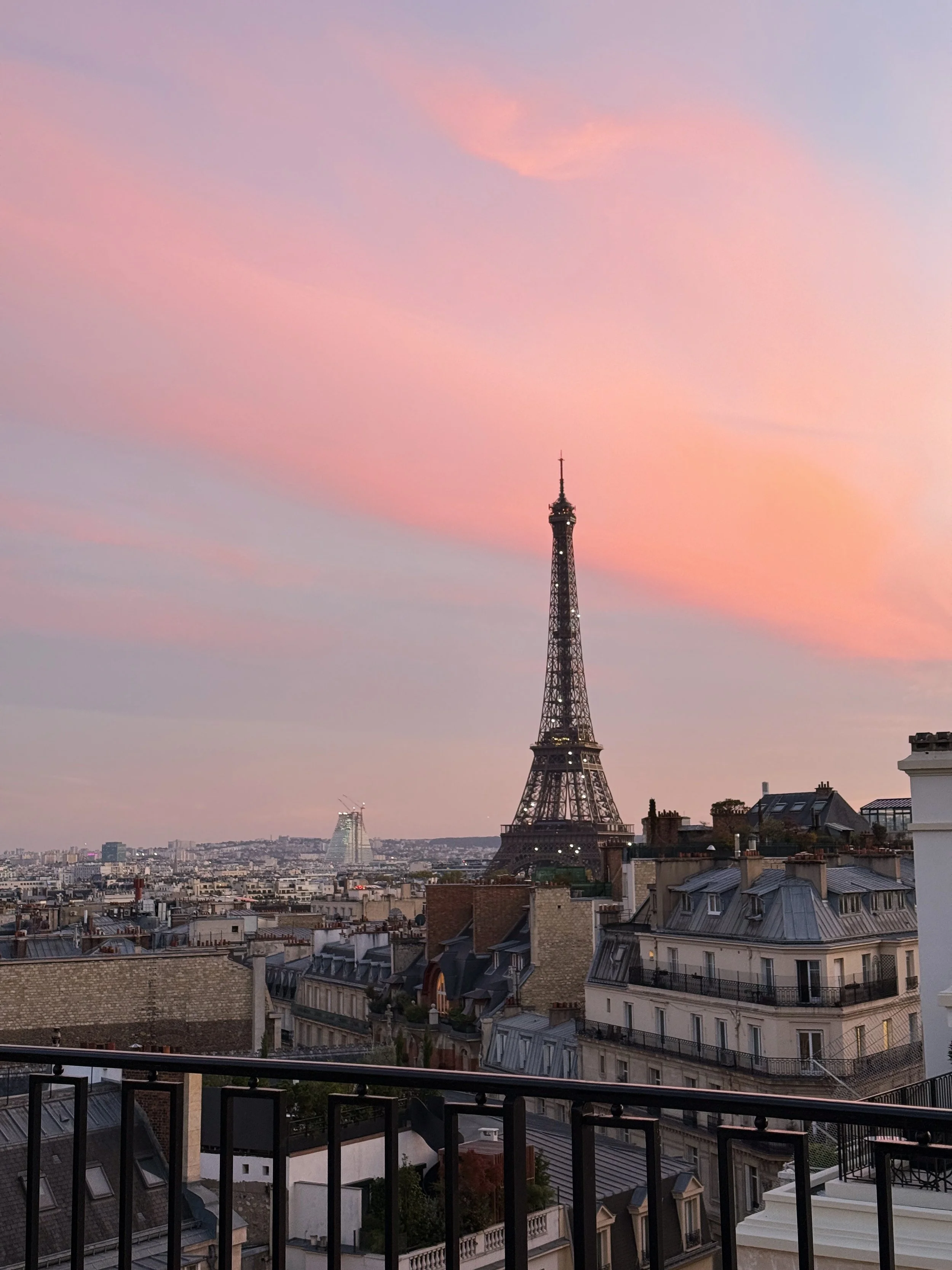Paris skyline with the Eiffel Tower at sunset, showing pink and purple clouds in the sky and rooftops of neighboring buildings.