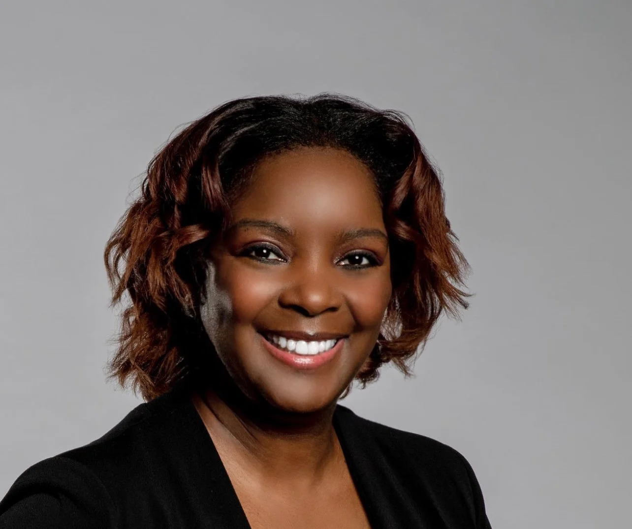 A portrait of an African American woman smiling, with shoulder-length wavy hair, wearing a black blazer, against a plain gray background.
