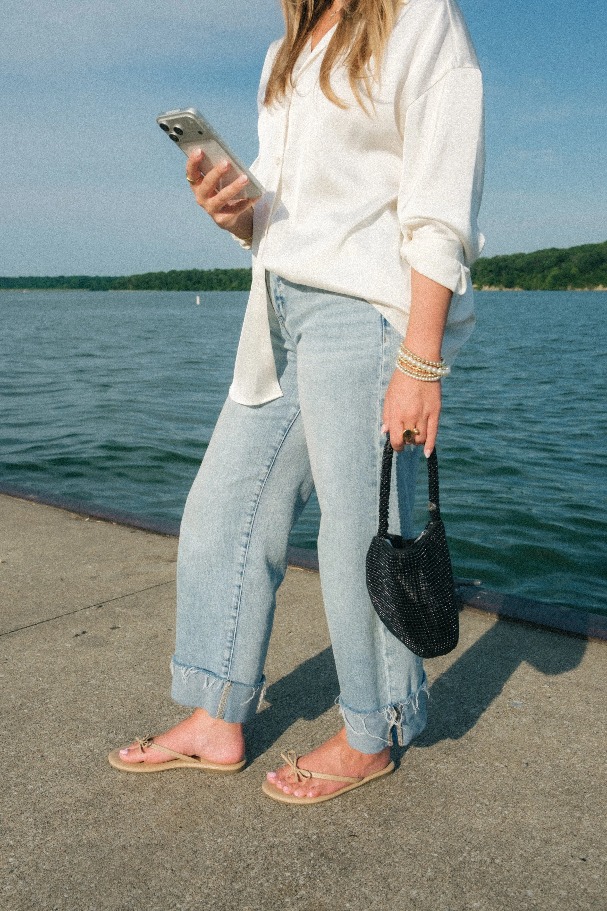A woman standing by a body of water, wearing a white shirt with rolled-up sleeves, light blue ripped jeans, and tan slide sandals with bows. She has pearl bracelets, a ring, and is holding a black woven handbag. She is looking at a smartphone in her hand, with her face not visible.