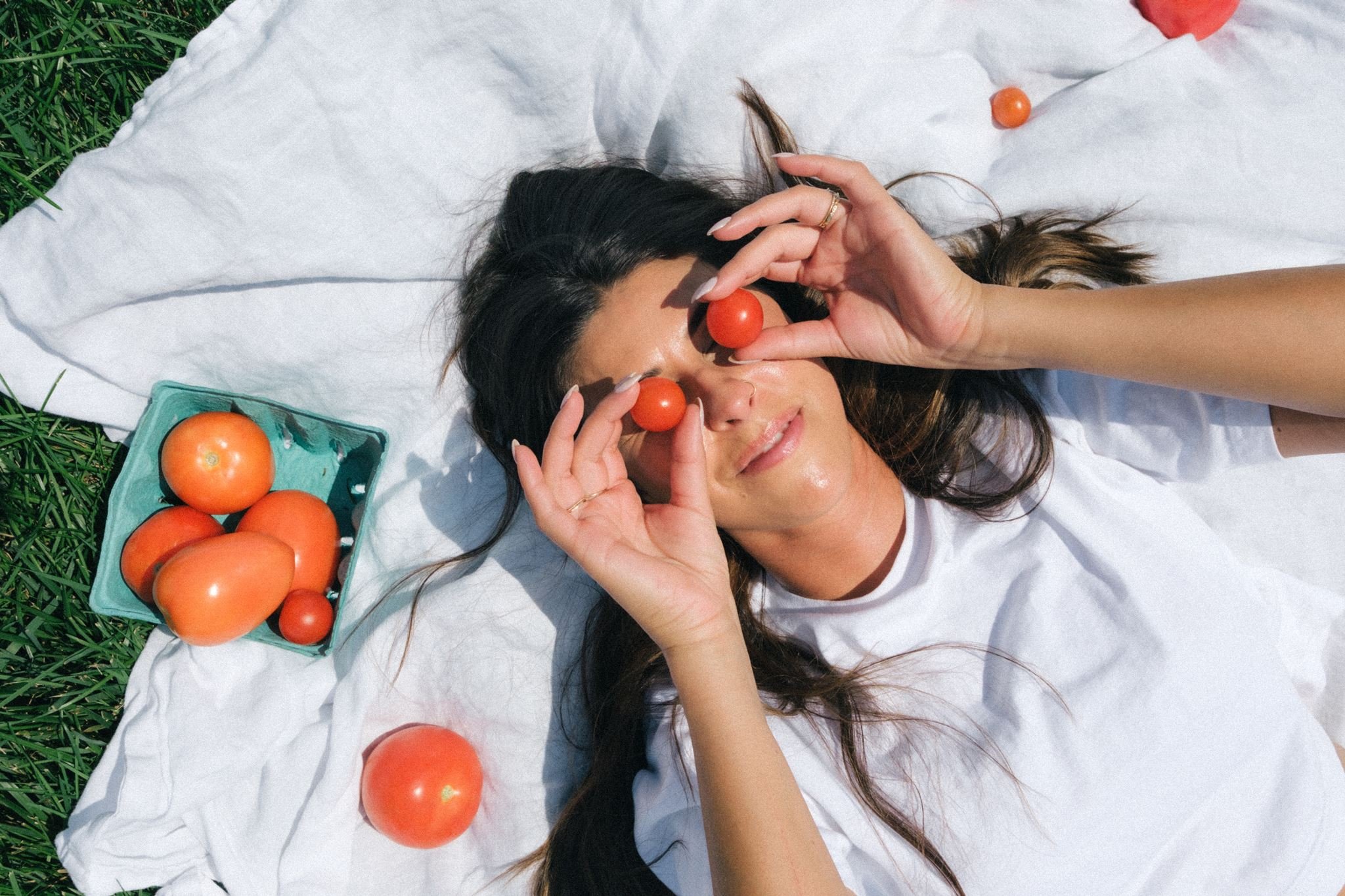 A woman laying on a blanket outside, holding tomatoes to her eyes for creative brand photos.