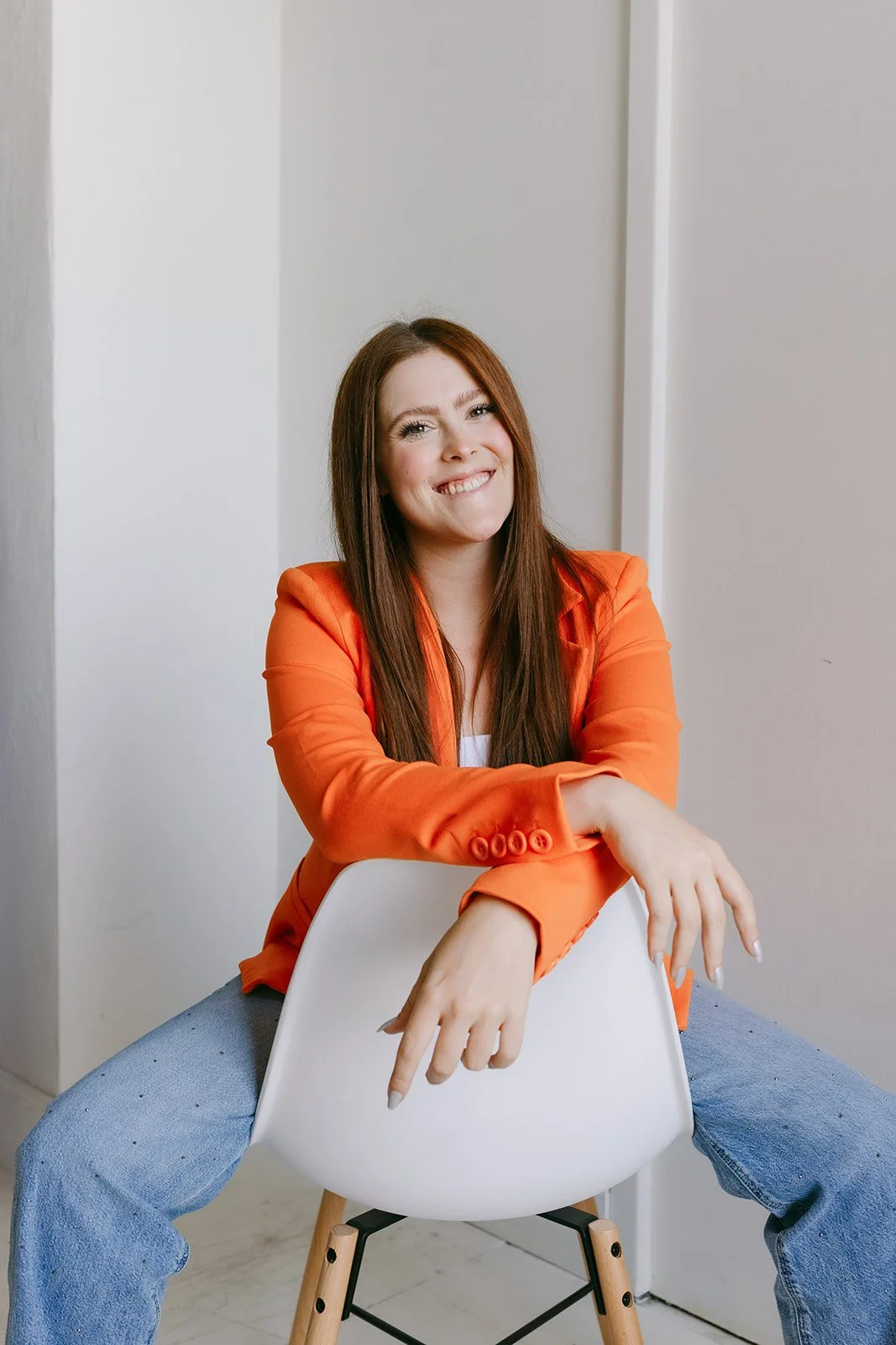  A woman sitting backwards in a chair wearing a bold orange blazer for business headshots.  