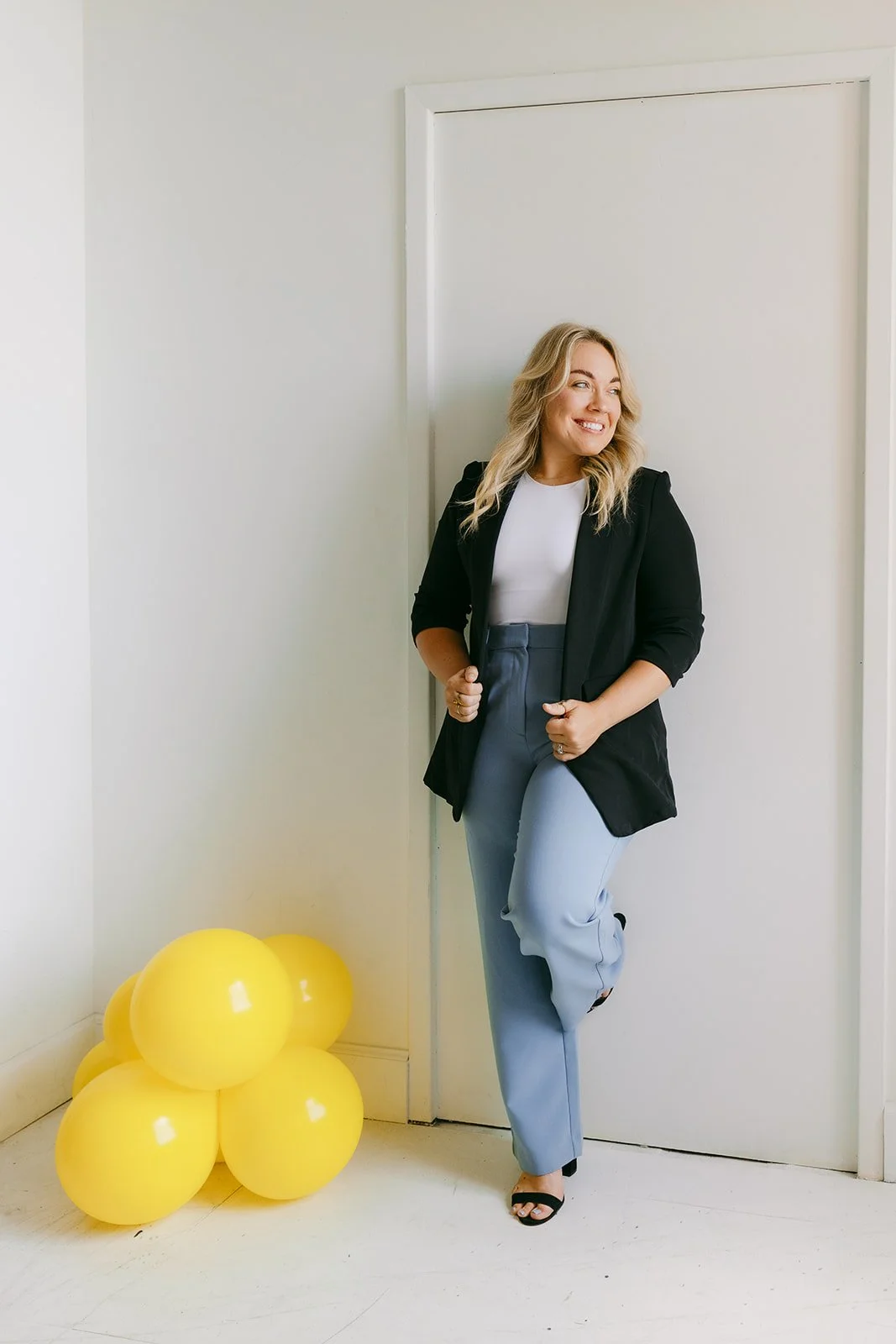 A woman leaning against a wall and looking to the side with yellow balloons on the ground.