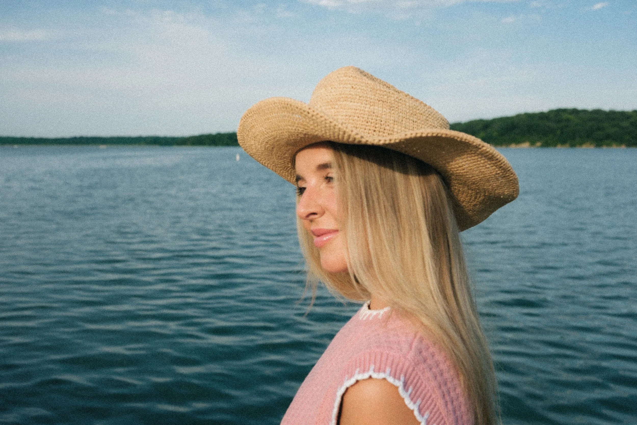 A woman wearing a wide-brimmed straw hat and a pink sweater with white scalloped edges, standing near water with a distant shoreline and green trees in the background.