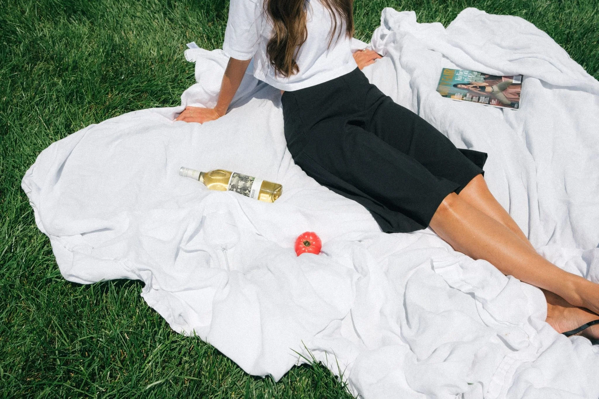 A business owner sitting in the grass on a white blanket with photoshoot prop ideas such as a magazine, wine, and tomato.