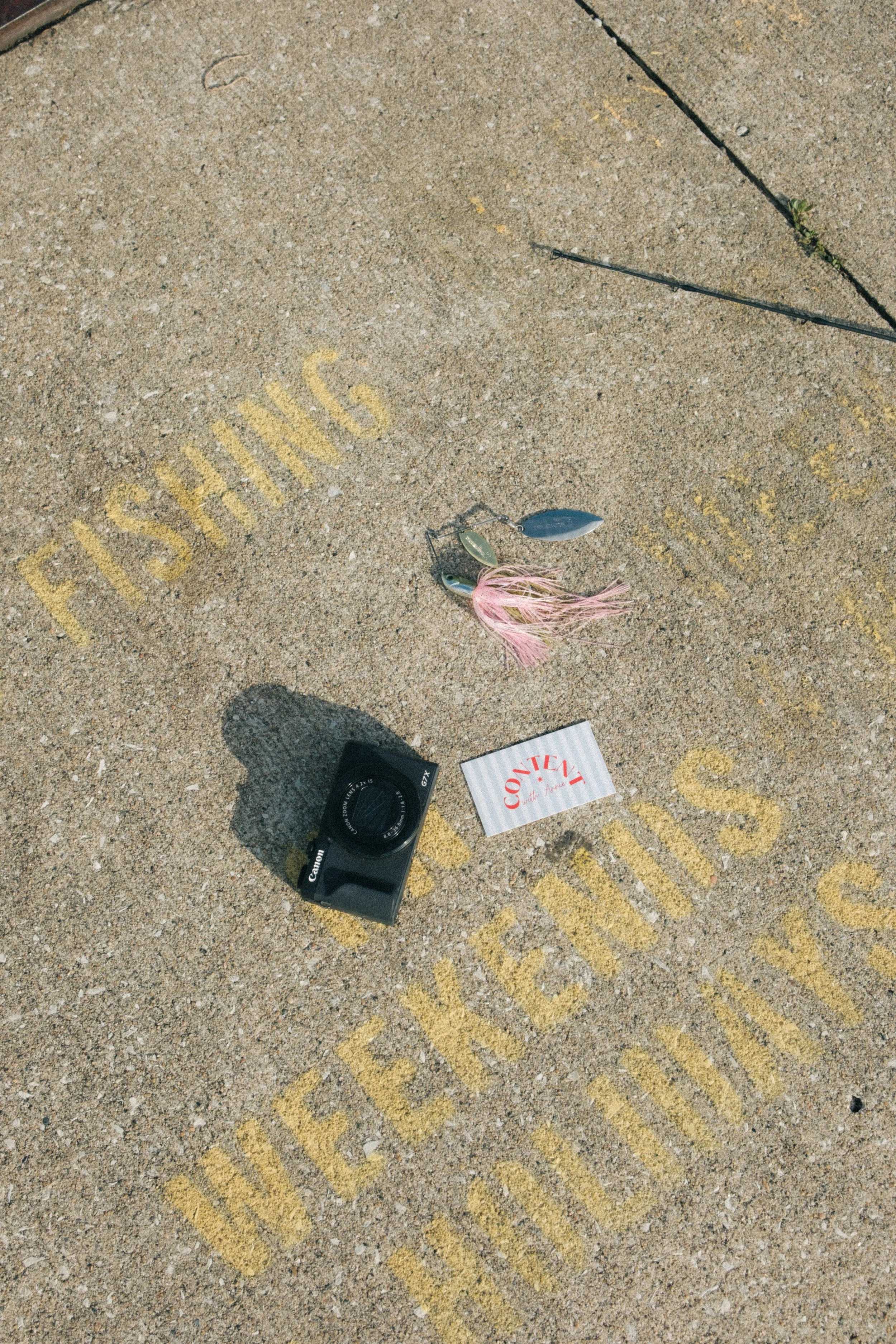 A concrete sidewalk with yellow painted words that say 'DO NOT STOP' and a golf lure with a pink feather and two metal blades, along with a camera and a white paper.