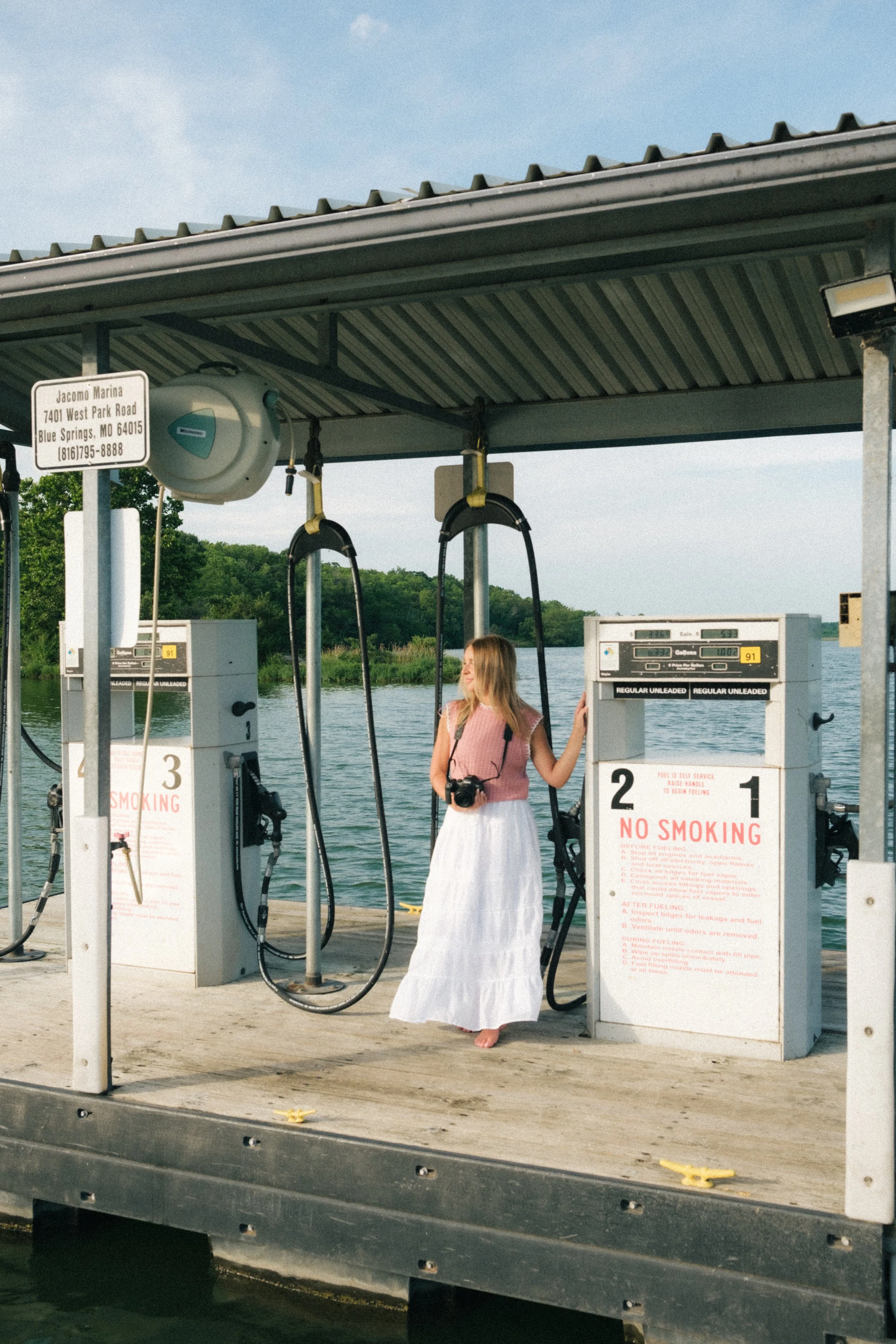 A woman in a white skirt and pink top standing barefoot on a dock at a gas station by the water, holding a camera with a pink strap, surrounded by fuel pumps under a metal roof, with a sign showing the station's address in Blue Springs, Missouri and a water body in the background.