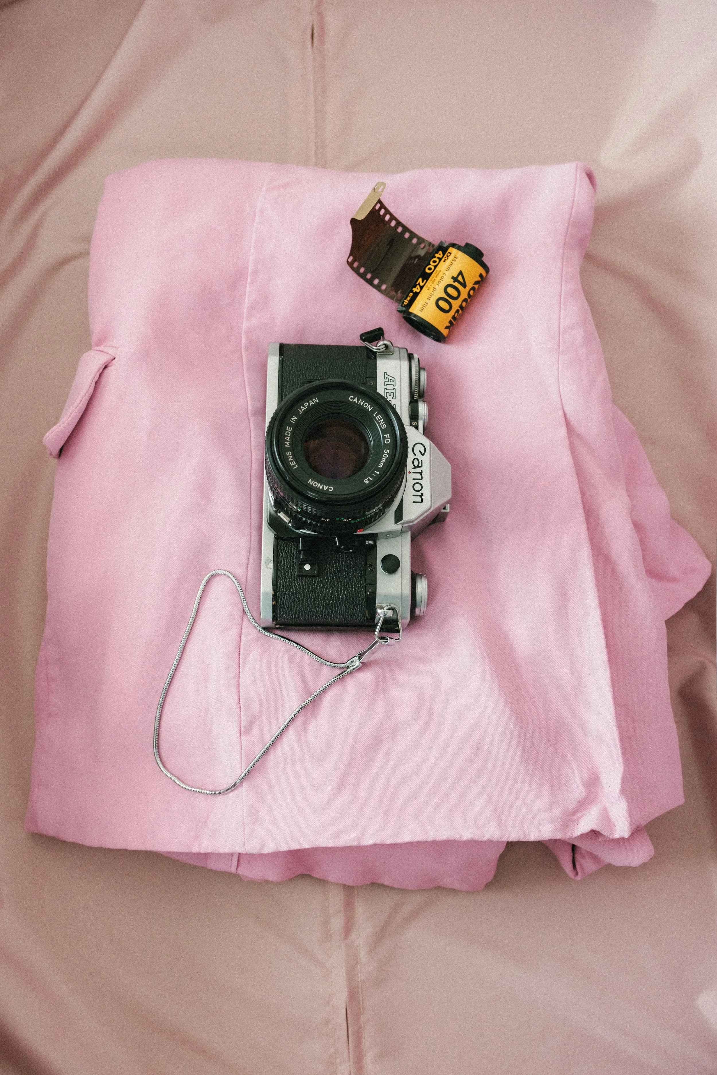 A vintage Canon film camera with black and silver body and attached lens, resting on a pink fabric pillow, with a roll of Kodak 400 film and a strip of exposed film above it.