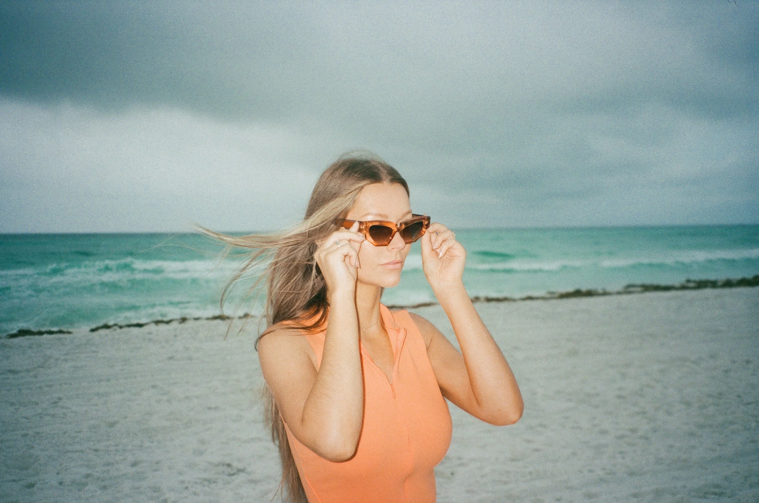 Woman with long hair adjusting sunglasses on a beach with ocean waves and cloudy sky.