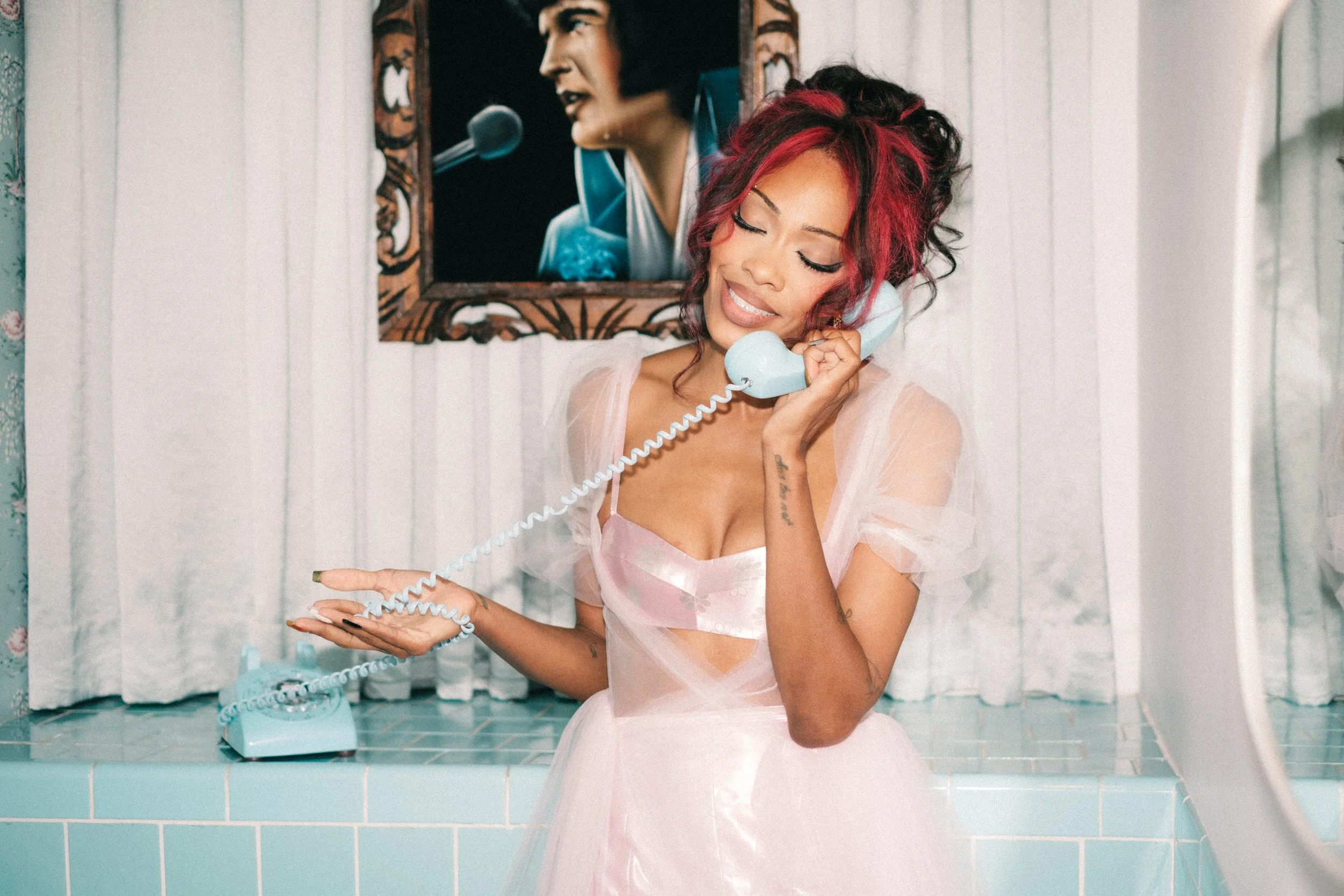 A young woman with red and black curly hair and tattoos, wearing a pink dress, is smiling with eyes closed while holding a vintage light blue telephone receiver to her ear. She is sitting in front of a white tiled wall with a framed picture of a woman singing into a microphone in the background.