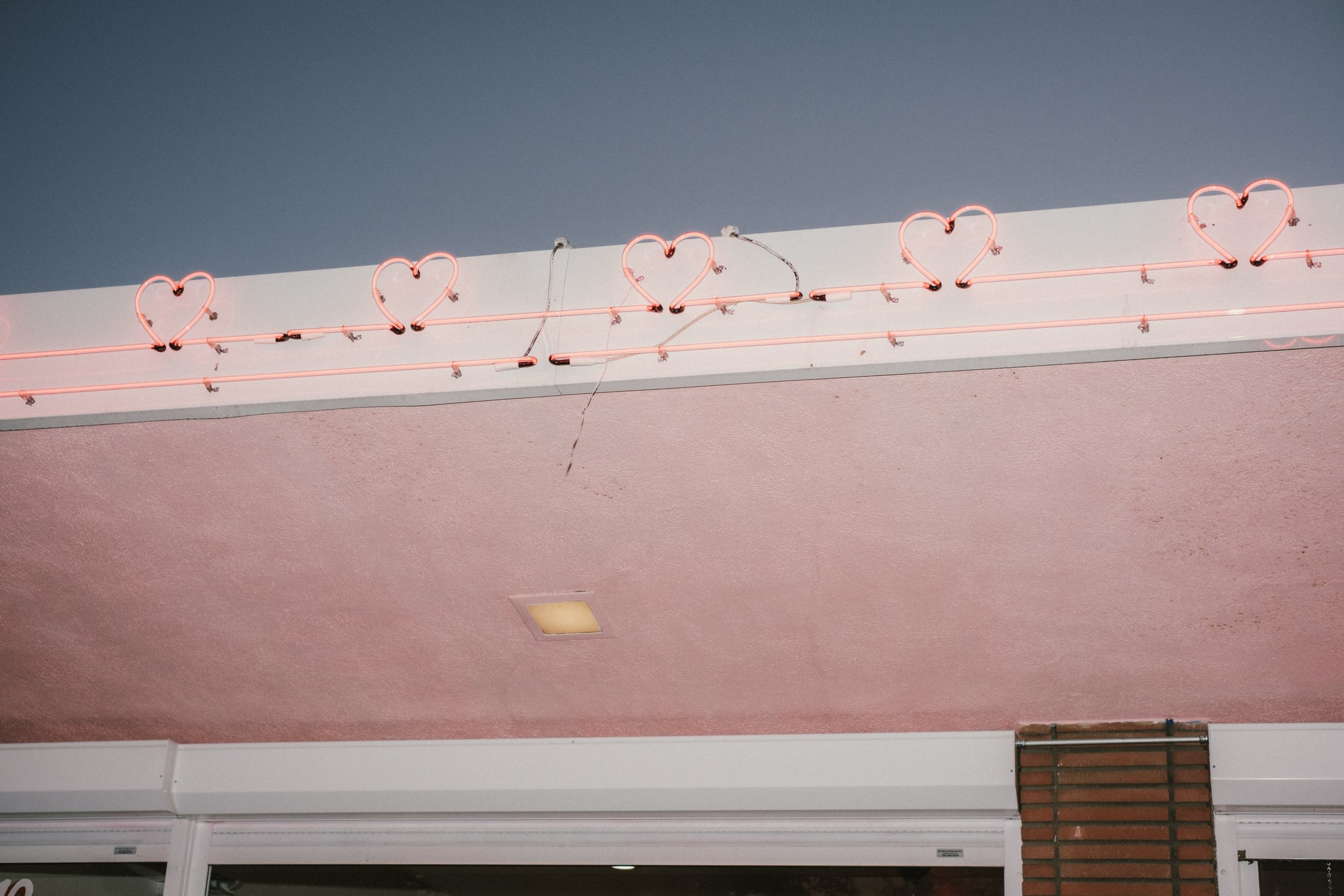 Pink neon sign with hearts mounted above shop entrance, pink ceiling with light fixture, brick column at the corner.