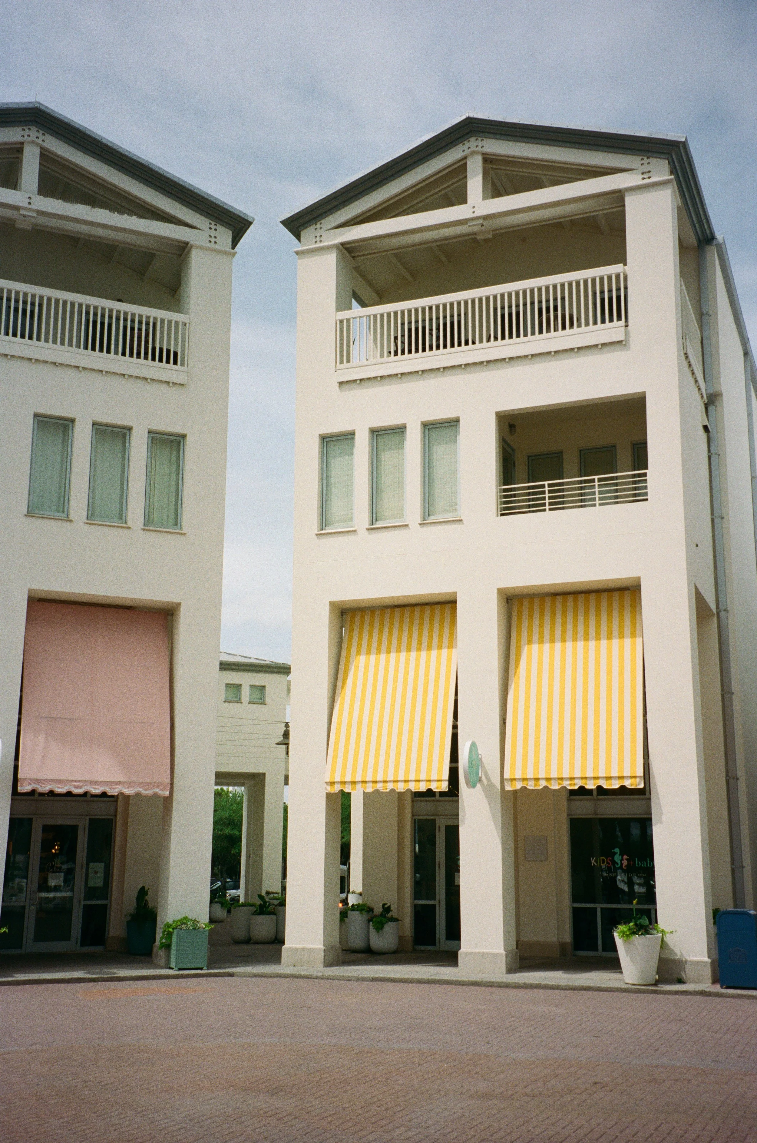 Two white buildings with open balconies, large windows, and yellow and white striped awnings. The ground level features glass doors and storefronts.