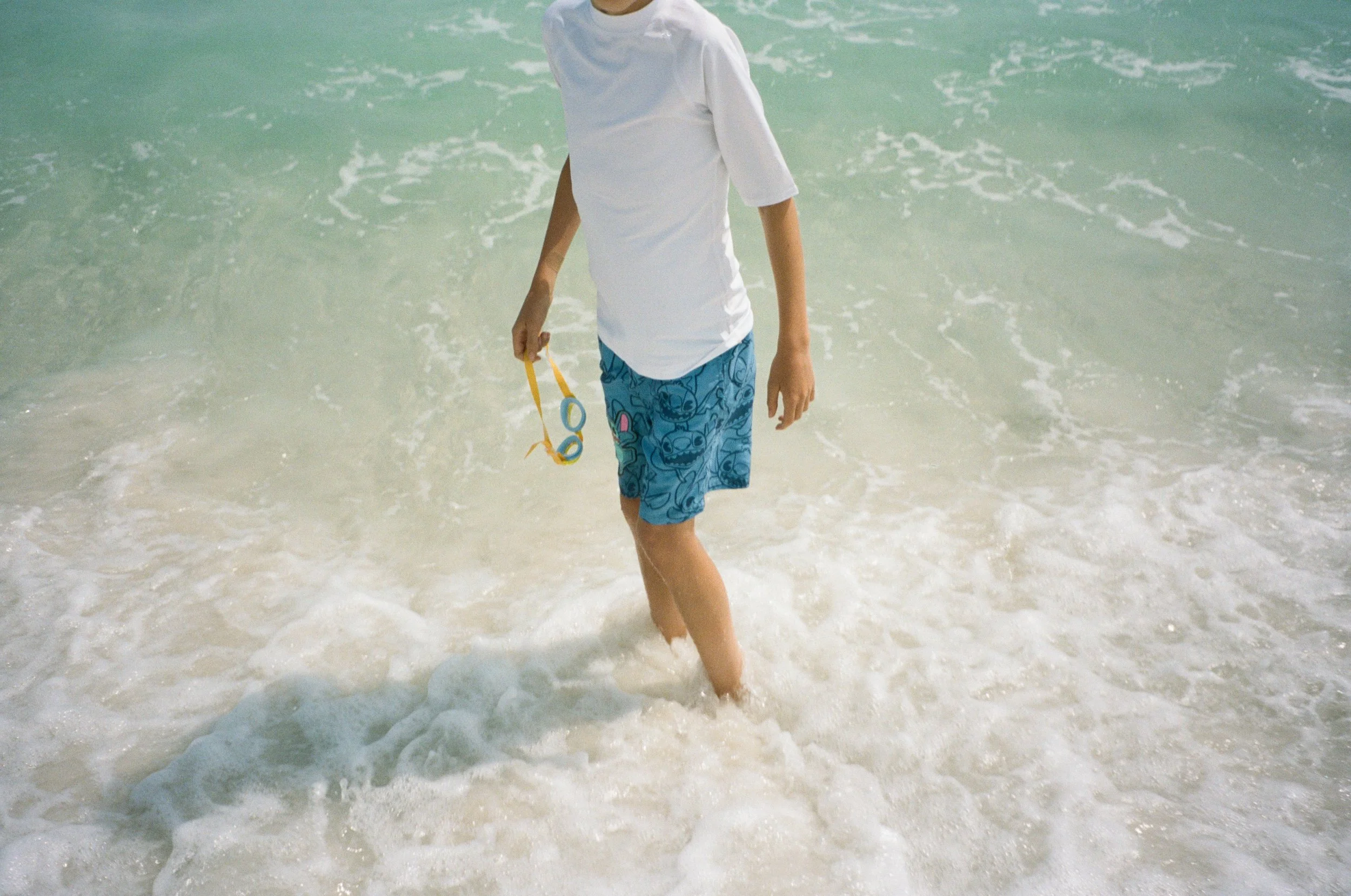 A person walking in the shallow ocean water, holding goggles in one hand, wearing a white t-shirt and blue shorts with cartoon prints.