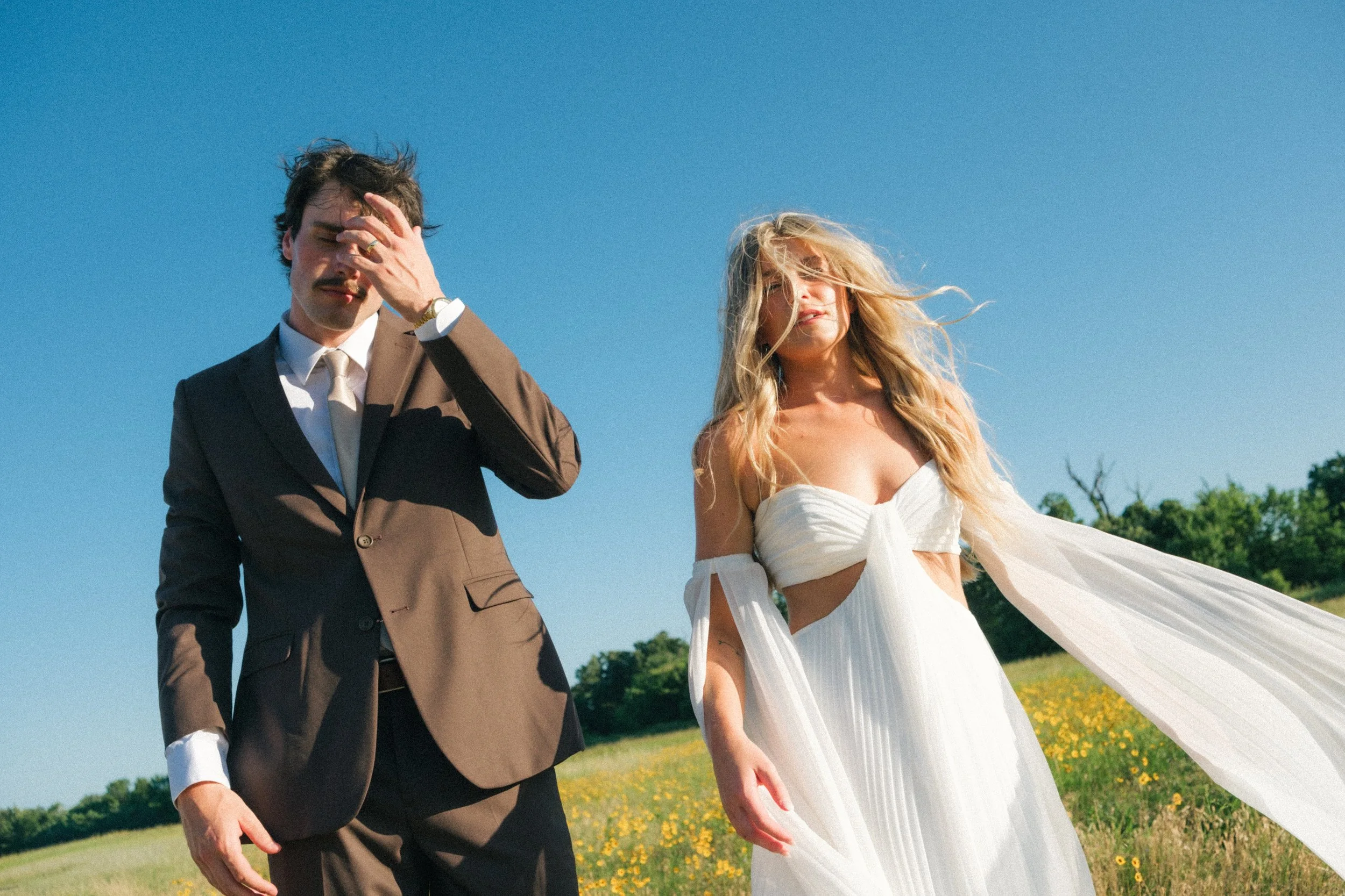 A man in a brown suit with a white shirt and tie covering his face with his hand, standing next to a woman in a white dress outdoors on a sunny day. The woman has long blonde hair and is looking at the camera with wind-blown hair, standing in a field with yellow flowers and green trees in the background.