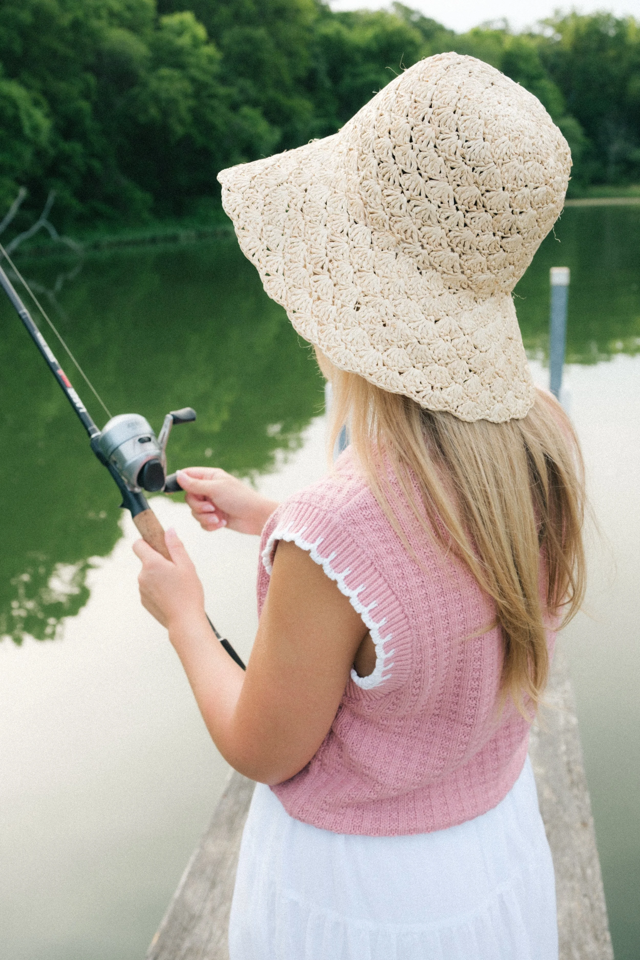 A girl fishing by a lake wearing a pink sleeveless sweater, white skirt, and large straw hat, with trees reflected in the water.