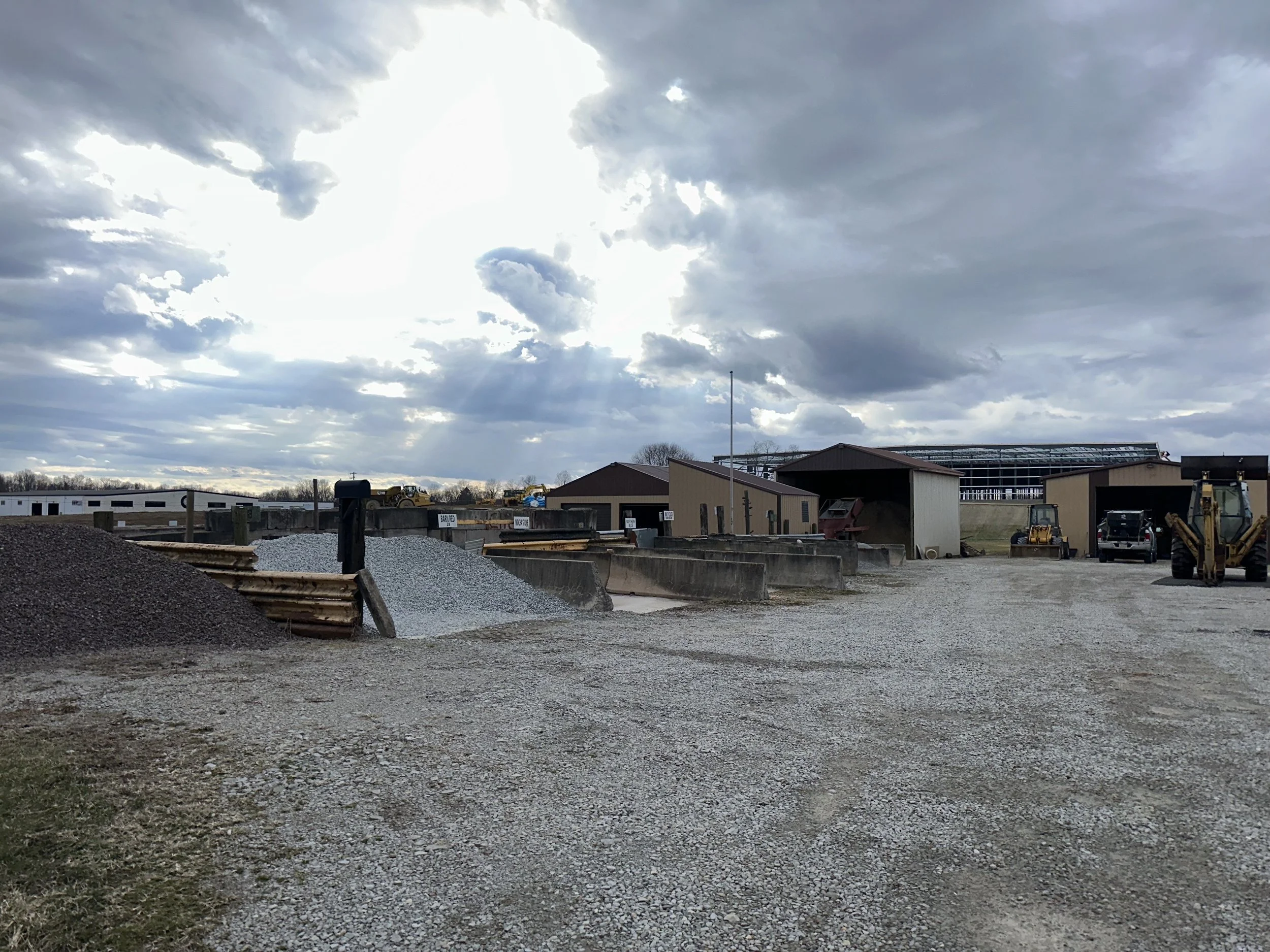 Industrial yard with gravel piles, storage buildings, and construction equipment under a cloudy sky.