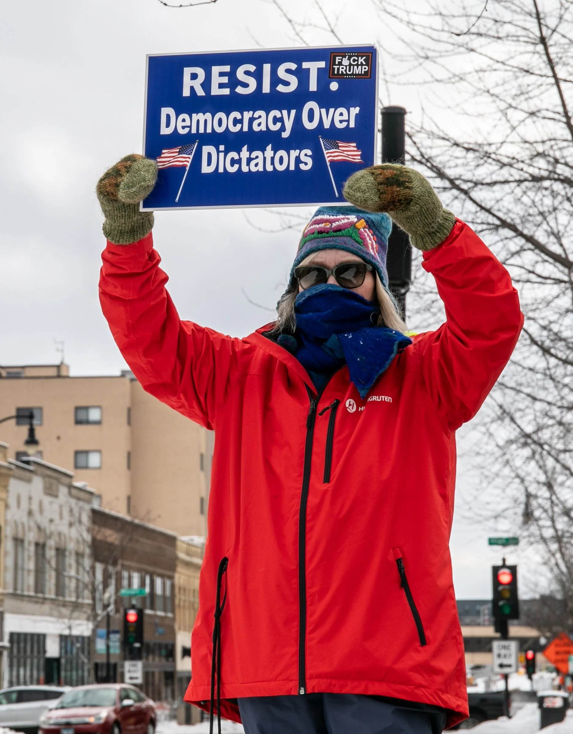 Person in red jacket holding a sign that says "RESIST. Democracy Over Dictators" with American flags and a sticker that says "FUCK TRUMP", standing outdoors in a city with snow on the ground, overcast sky, leafless trees, and surrounding buildings.