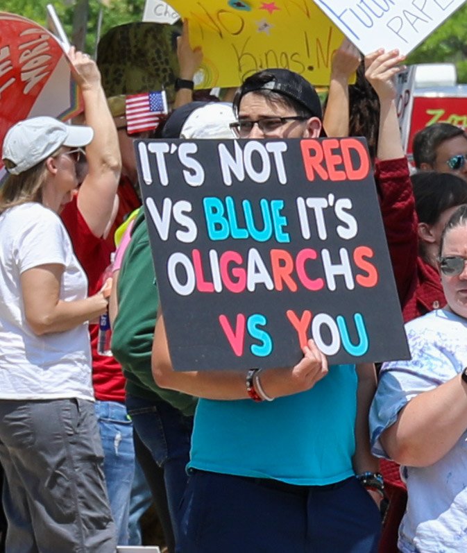 A person in a blue shirt holding a protest sign that reads, 'It's not red vs blue it's oligarchs vs you,' amidst a crowd of protesters with various signs.