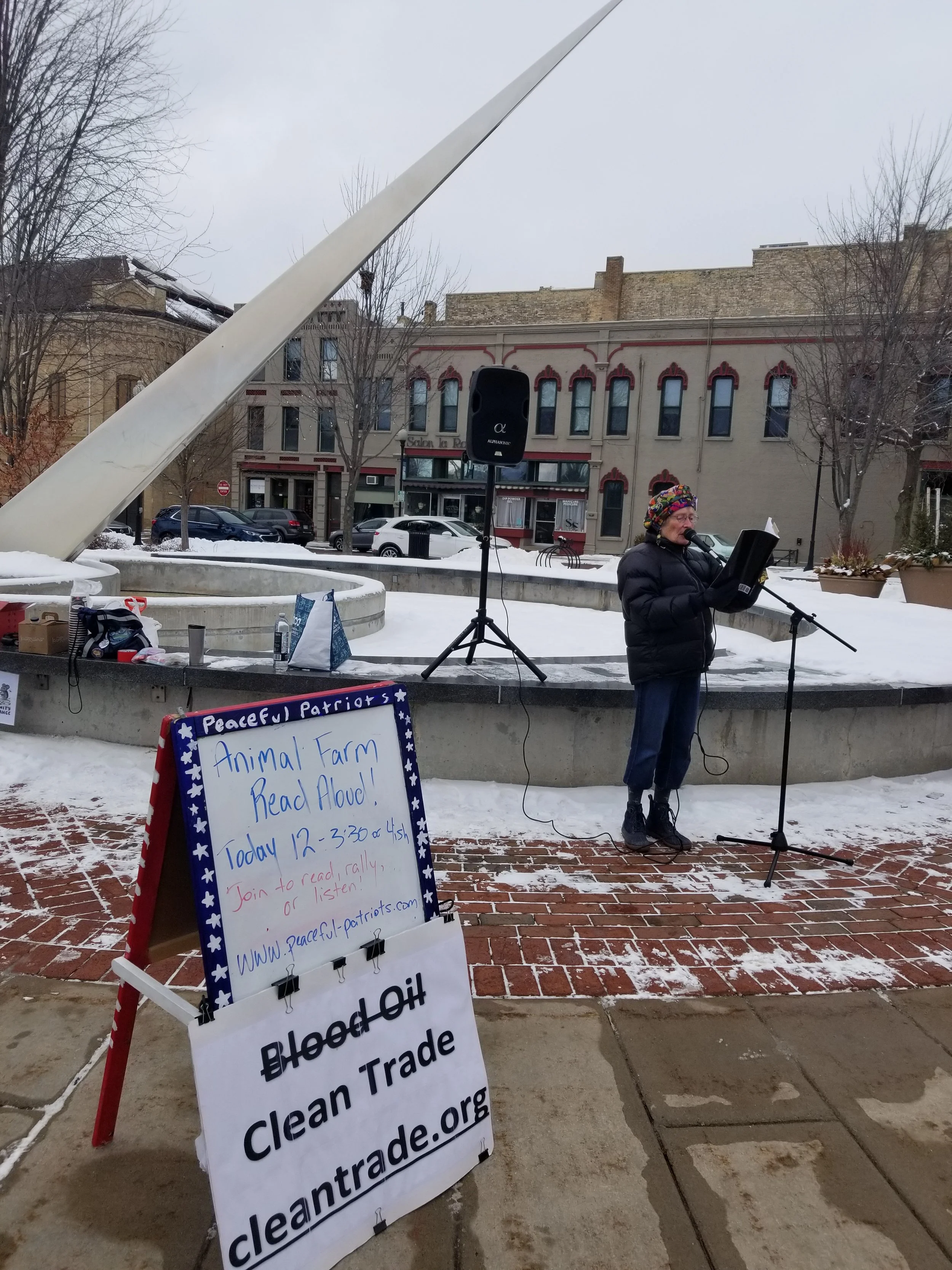 A woman reading aloud into a microphone outdoors in a snowy town square with a bronze sculpture, a sign promoting Animal Farm read aloud event, and a sign for Blood of Blood Clean Trade.