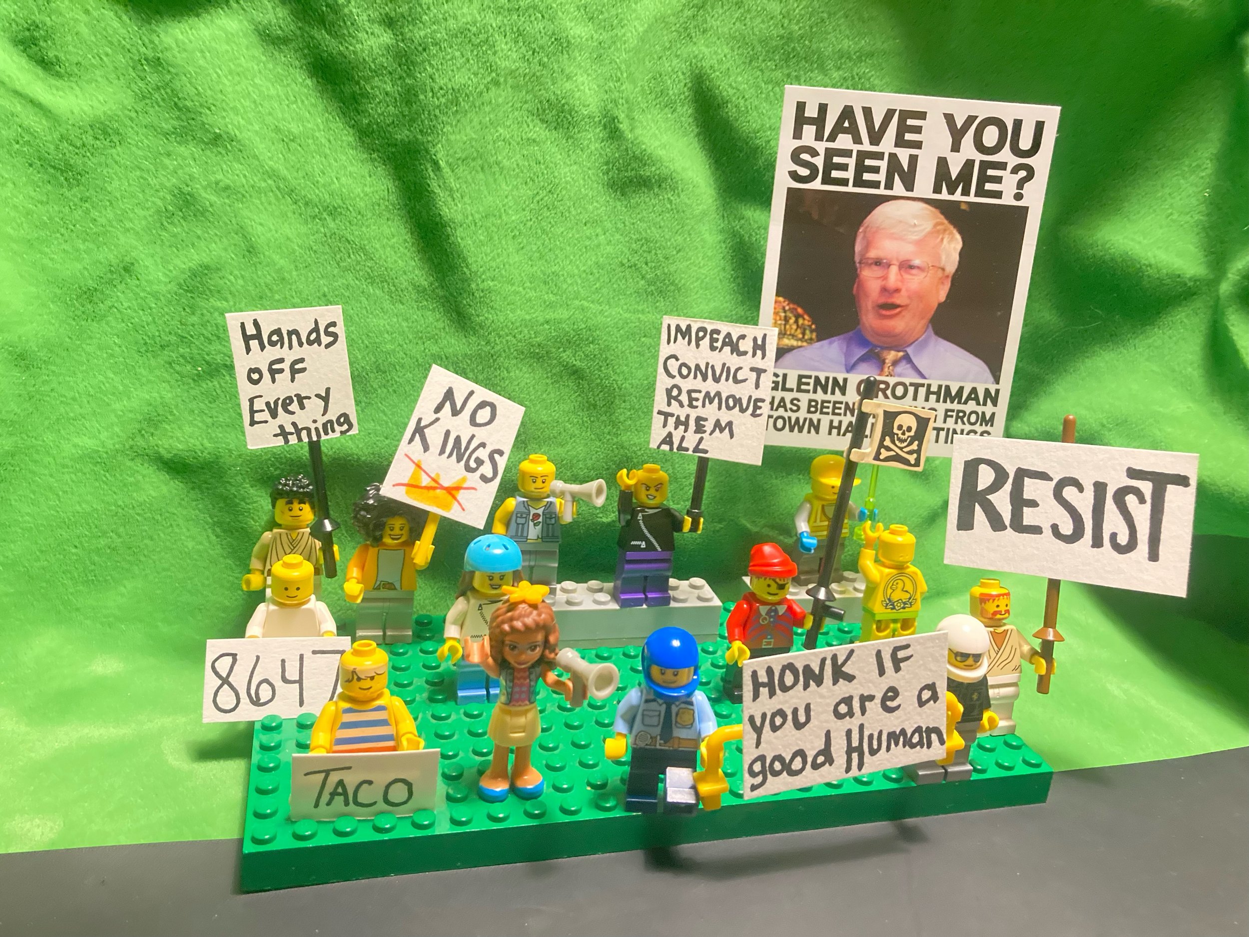 Lego figures holding protest signs on a green background with a large photo of a man in the background. Signs include messages like 'HANDS OFF EVERYTHING,' 'NO KINGS,' 'HAVE YOU SEEN ME?,' 'RESIST,' and 'HONK IF YOU ARE A GOOD HUMAN.'
