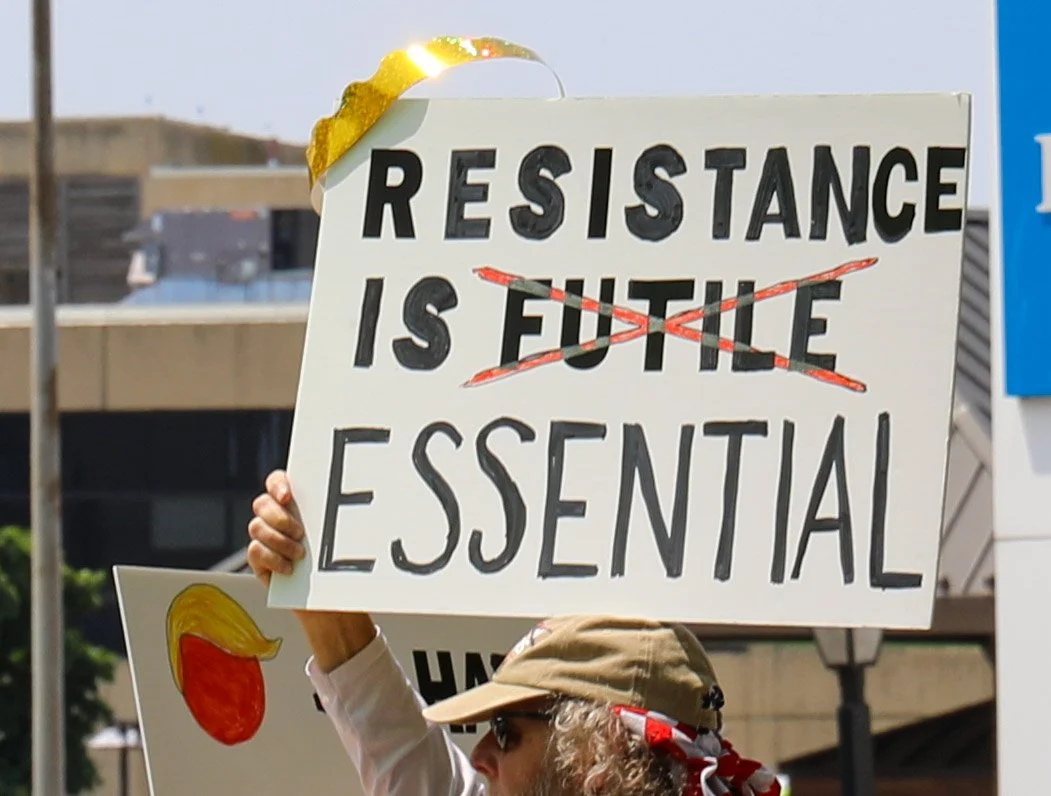 A protestor holding a sign that reads 'RESISTANCE IS FUTILE' with 'FUTILE' crossed out and replaced with 'ESSENTIAL'; other protesters are partially visible in the background.