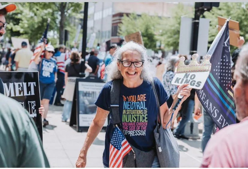 Smiling woman at a protest holding a sign that says 'No Kings' and a small American flag, surrounded by other protesters with signs and flags.