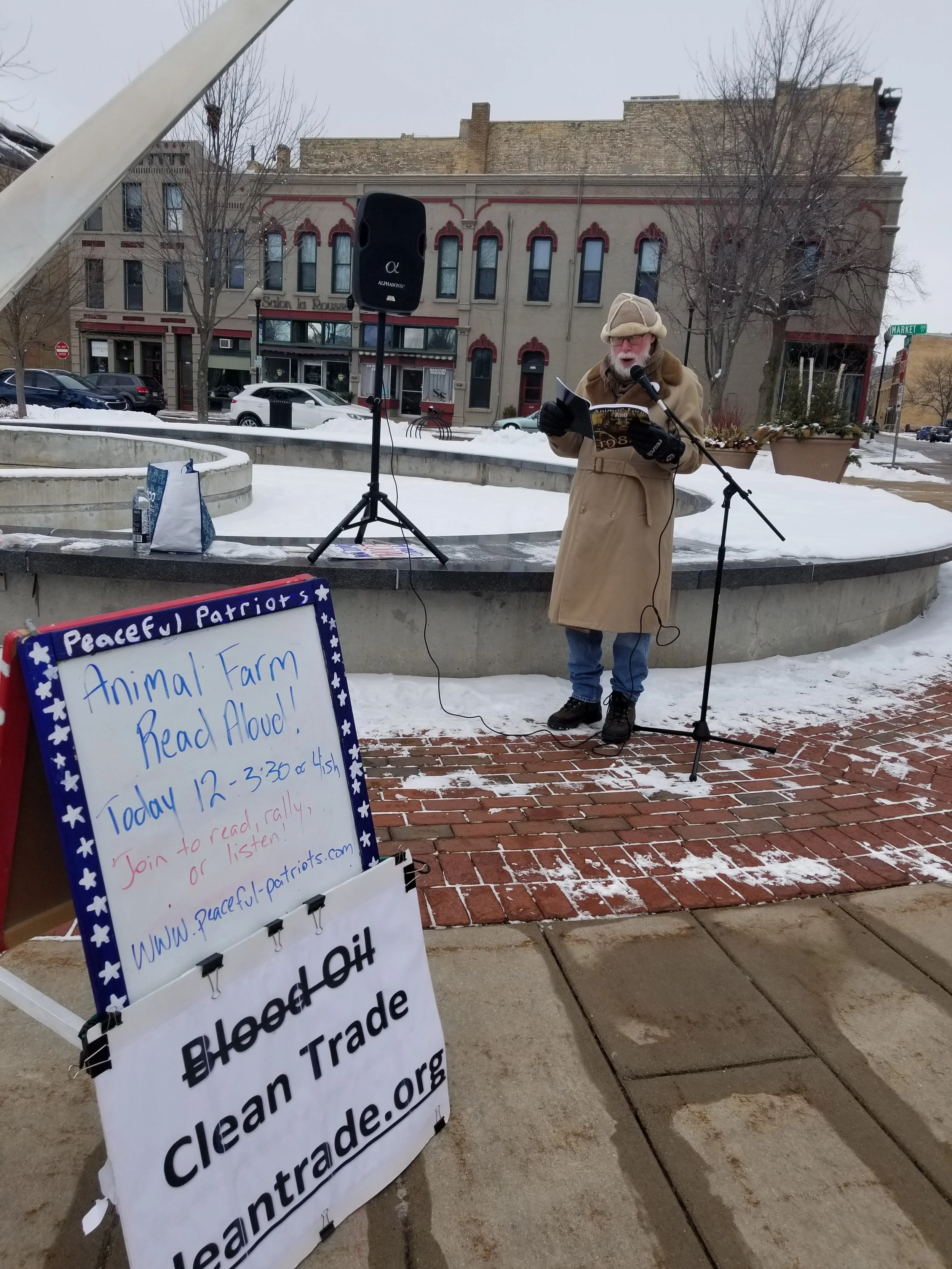 A man dressed in a beige coat, hat, and gloves is speaking into a microphone outdoors, reading from a paper. There is a sign promoting a political event about animal farm read-aloud and clean trade. Snow is on the ground, and buildings and trees are 