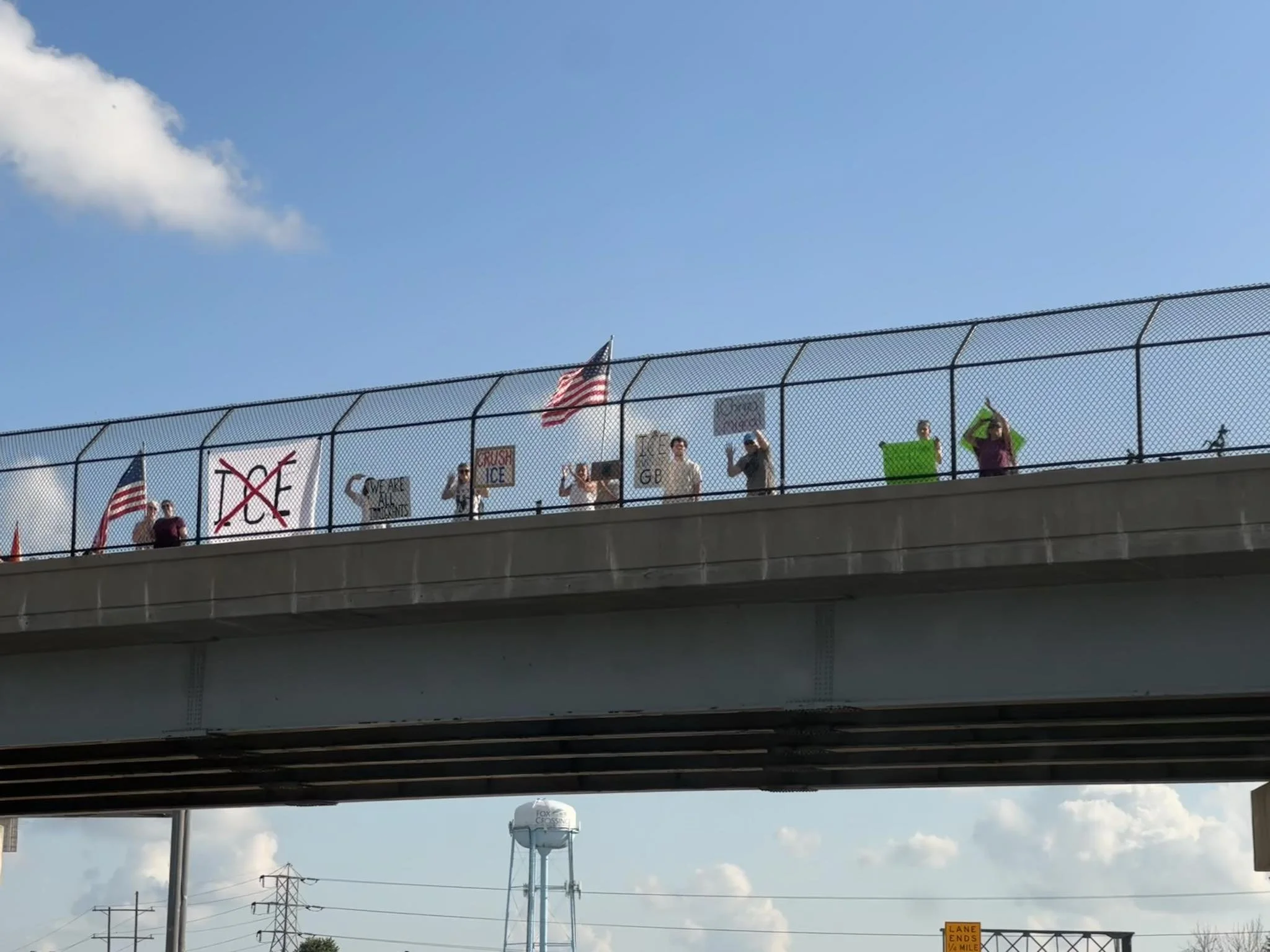 People standing on a bridge holding signs and flags during a protest or demonstration on a sunny day.