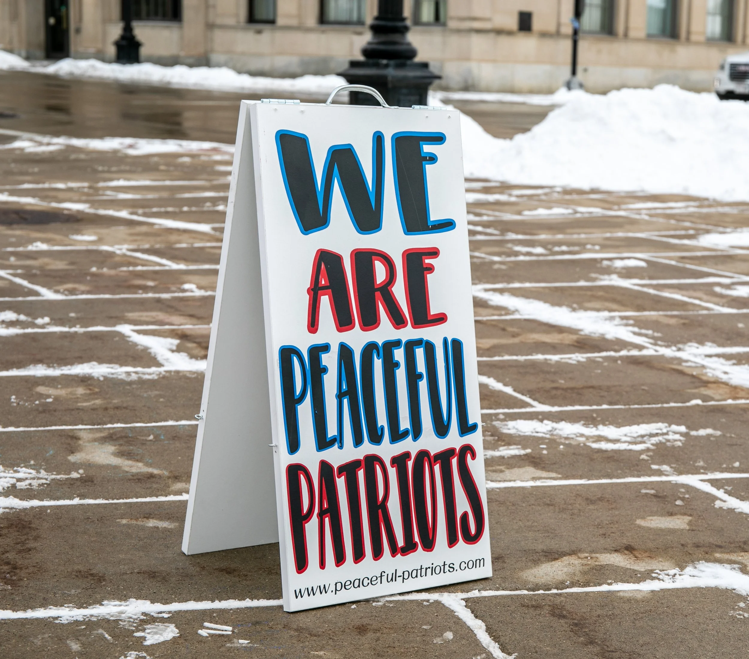 A white A-frame sidewalk sign with a message in bold blue, red, and black letters that reads 'We Are Peaceful Patriots' and a website URL at the bottom, placed on a wet, snow-covered pavement outdoors during winter.