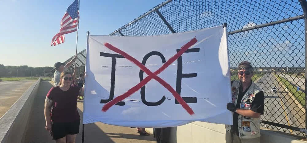 Two women standing on a bridge holding a large protest sign that has the word 'NOPE' crossed out with red lines. One woman is holding an American flag. The background shows a chain-link fence and the highway below.