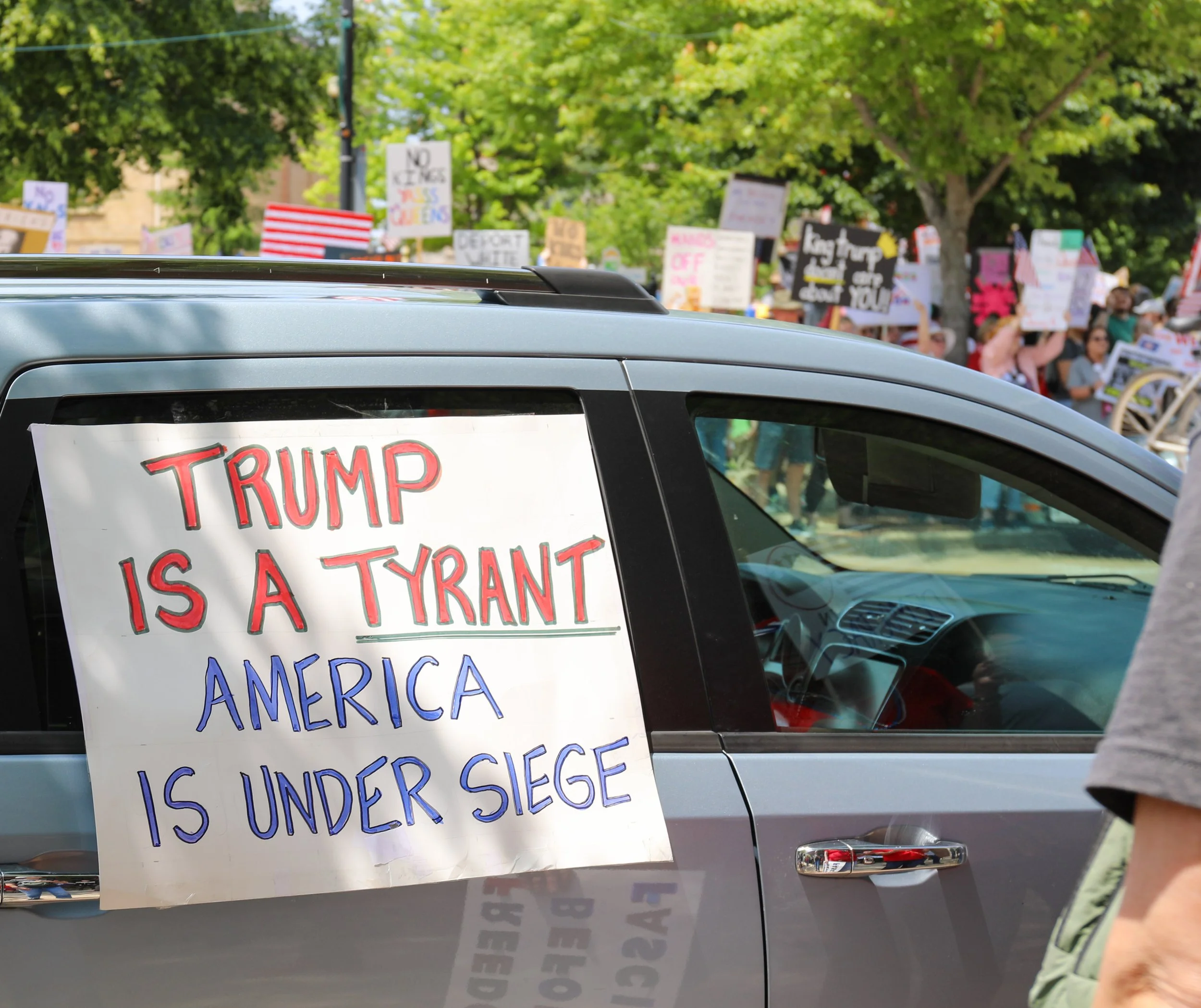 A sign on a car reading 'TRUMP IS A TYRANT. AMERICA IS UNDER SIEGE.' at a protest with a crowd of people holding signs in the background.