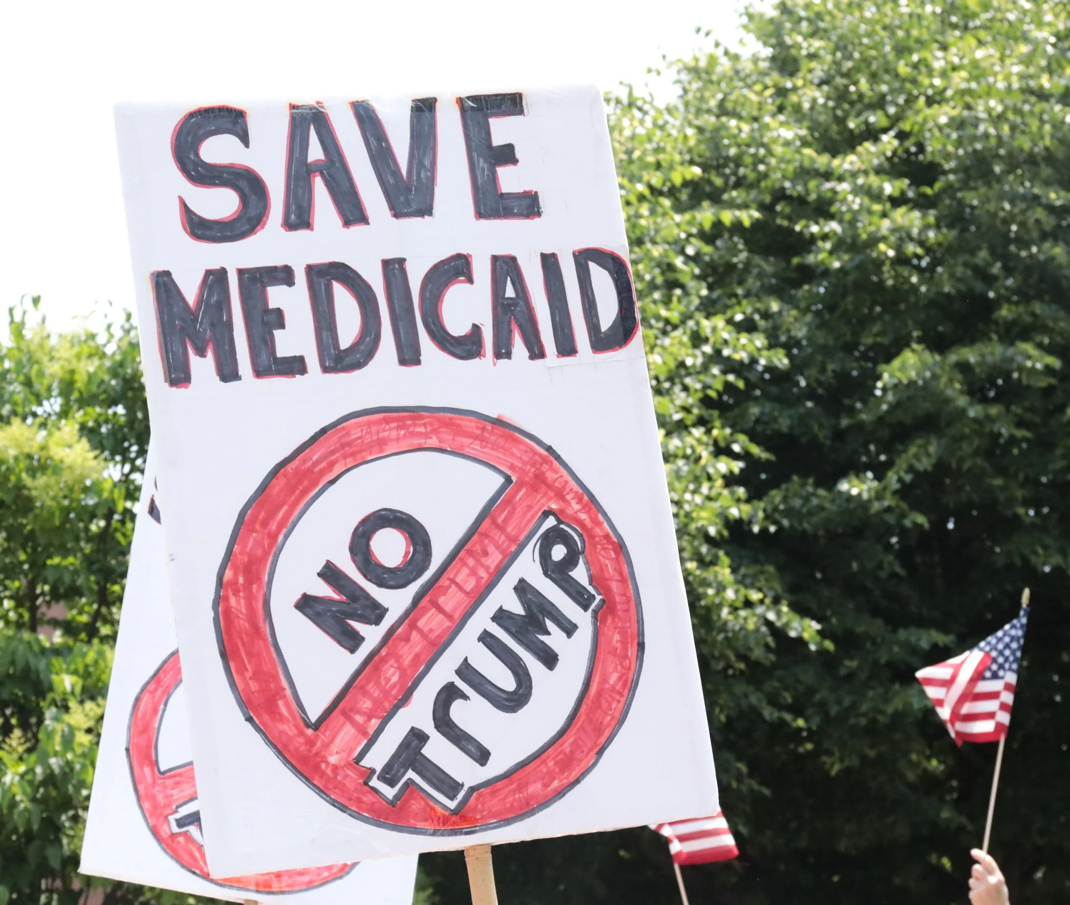 A protest sign that reads 'Save Medicaid' with a red circle and slash over the words 'No Trump', held among American flags during a demonstration.
