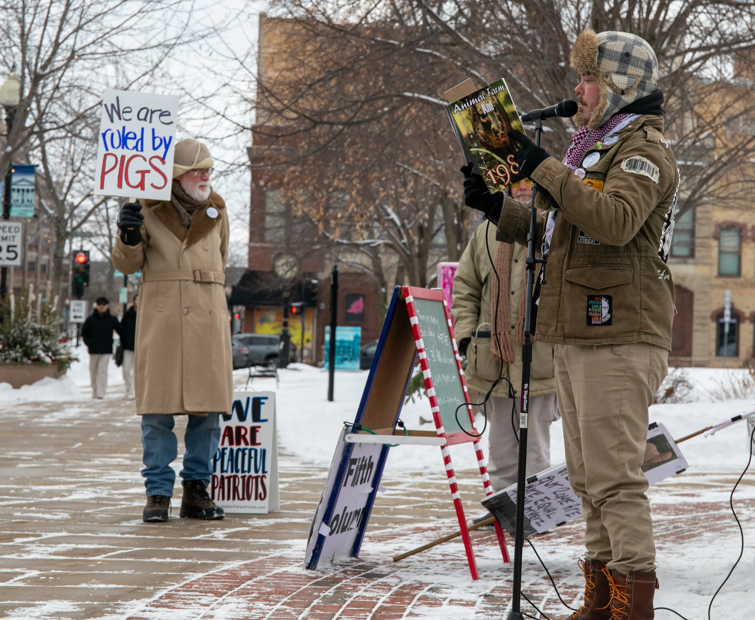 A man with a beard and glasses holding a sign that says "We are ruled by pigs" at a winter outdoor protest, with trees and buildings in the background.