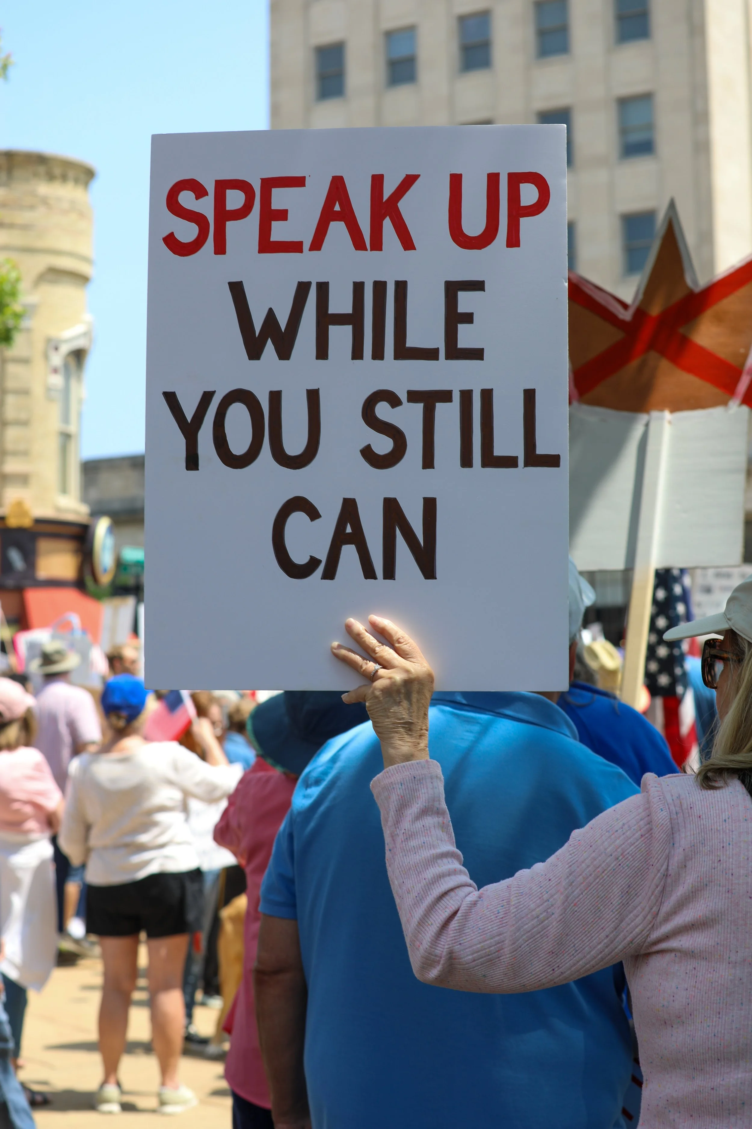 A person holding a sign that says 'SPEAK UP WHILE YOU STILL CAN' during a protest or rally, with a crowd of people and banners in the background.