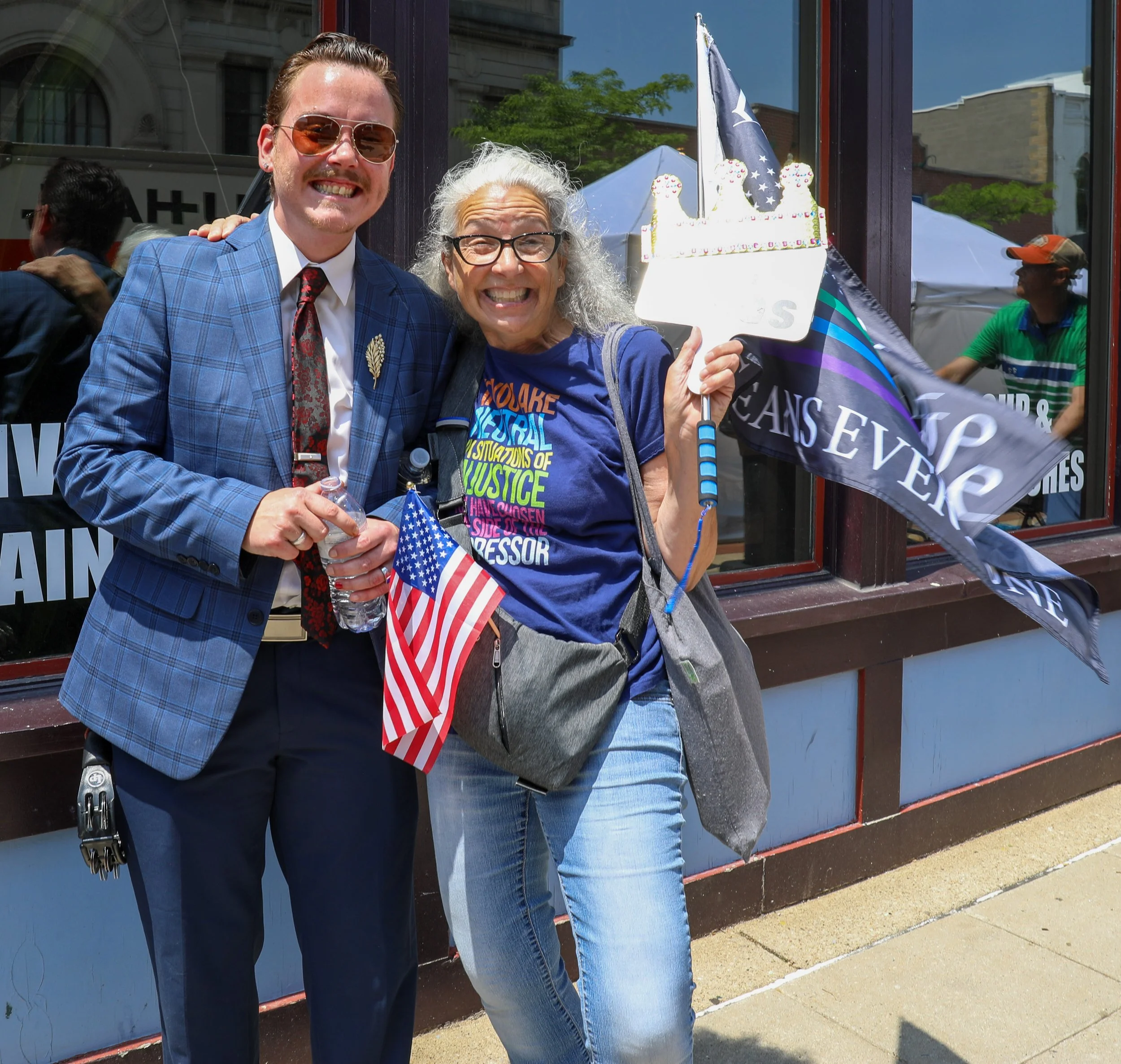 Two smiling women standing outside a building, holding American flags and protest signs, celebrating at a public event or rally on a sunny day.