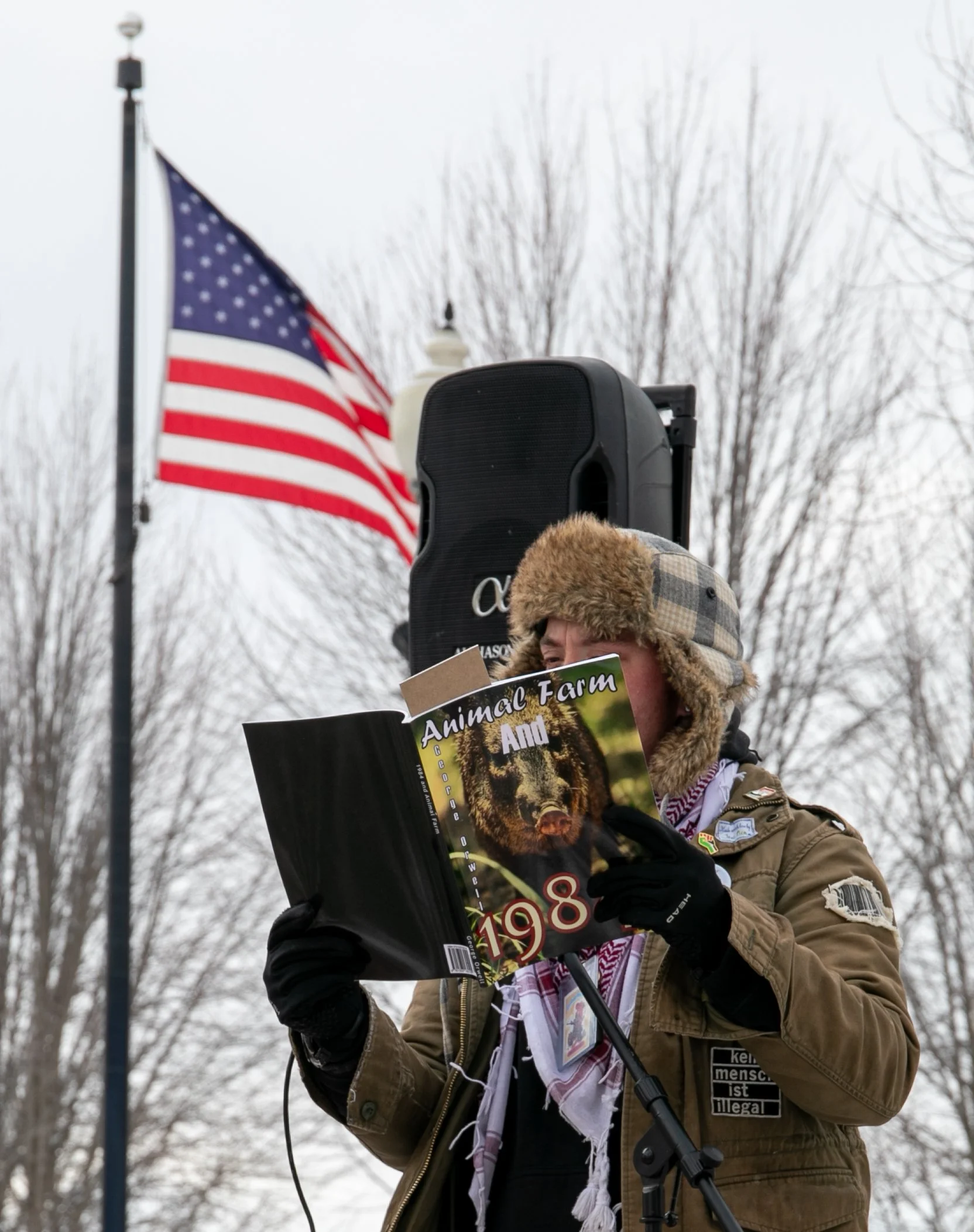 A woman wearing a fur-lined hat and brown jacket reading a magazine titled 'Animal Farm And 1984' near a flagpole with the American flag, outdoor setting with leafless trees in the background.
