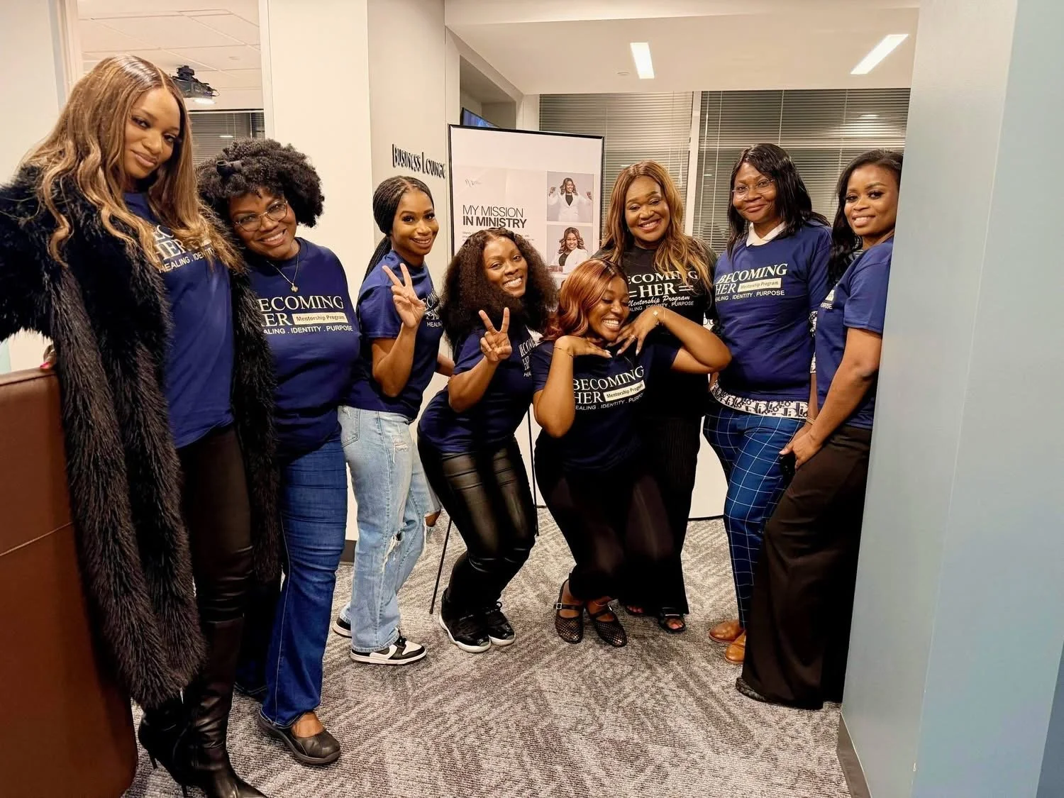 Group of women in matching blue shirts with the words 'Becoming Her' standing together in an office setting, smiling and posing for the photo.