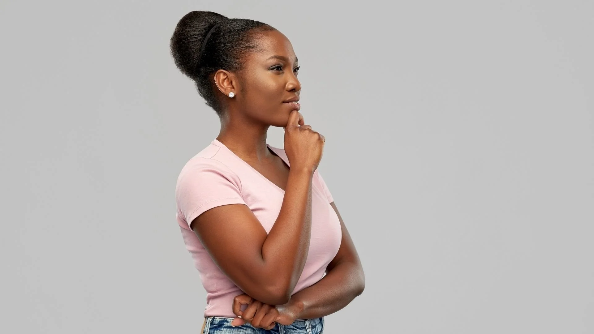 A young woman with dark hair styled in a bun, wearing a light pink t-shirt and jeans, standing against a plain gray background with her hand on her chin, appearing thoughtful.