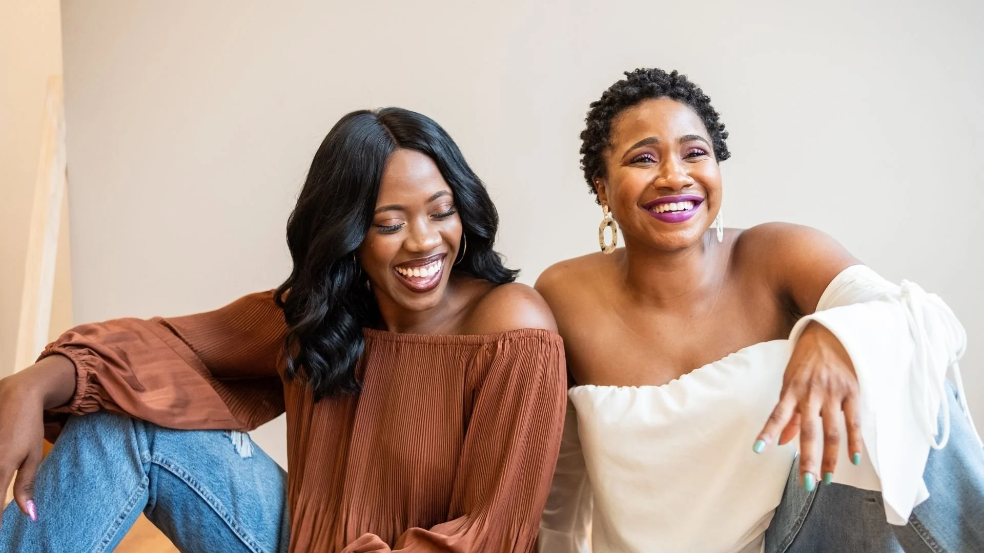 Two smiling women sit close together, one wearing a brown off-shoulder top and the other in a white off-shoulder top with earrings, against a plain background.