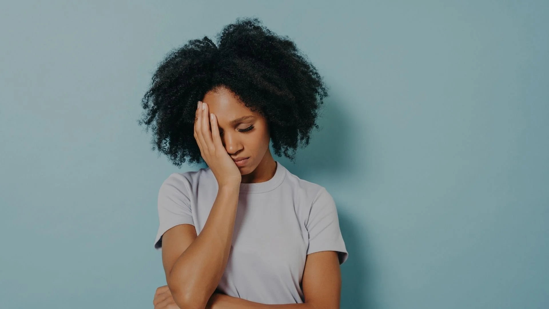 A young woman with curly black hair holding her forehead with one hand, looking down, with a concerned or worried expression, standing against a light blue background.