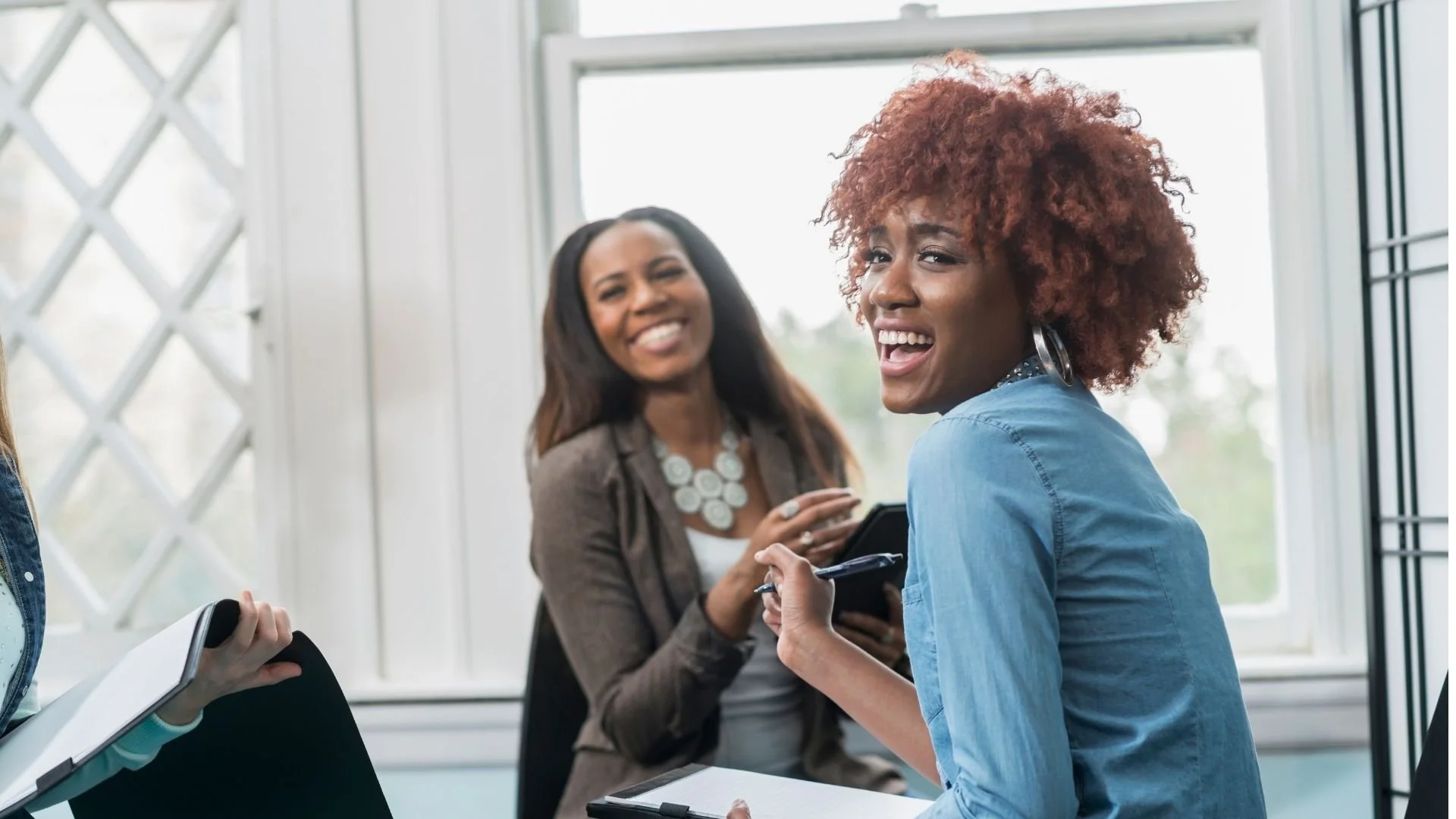 Two women smiling and sharing a laugh in an office setting; one holding a pen and notebook, the other with a tablet, near a large window.