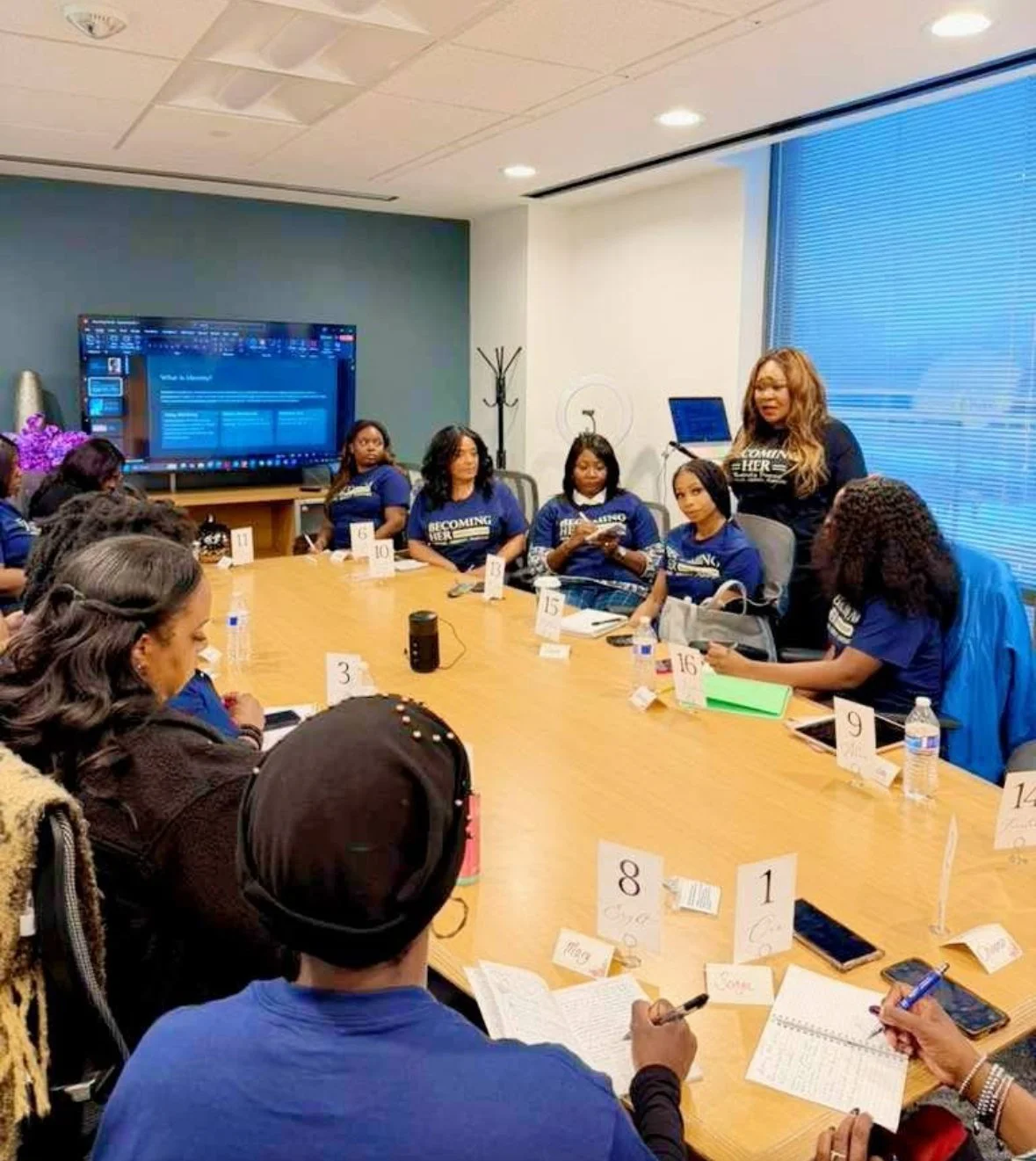 A group of women gathered around a conference table participating in a meeting or workshop. Some are taking notes, and one woman is standing and speaking. The room has a large screen, notebooks, water bottles, and name cards on the table.